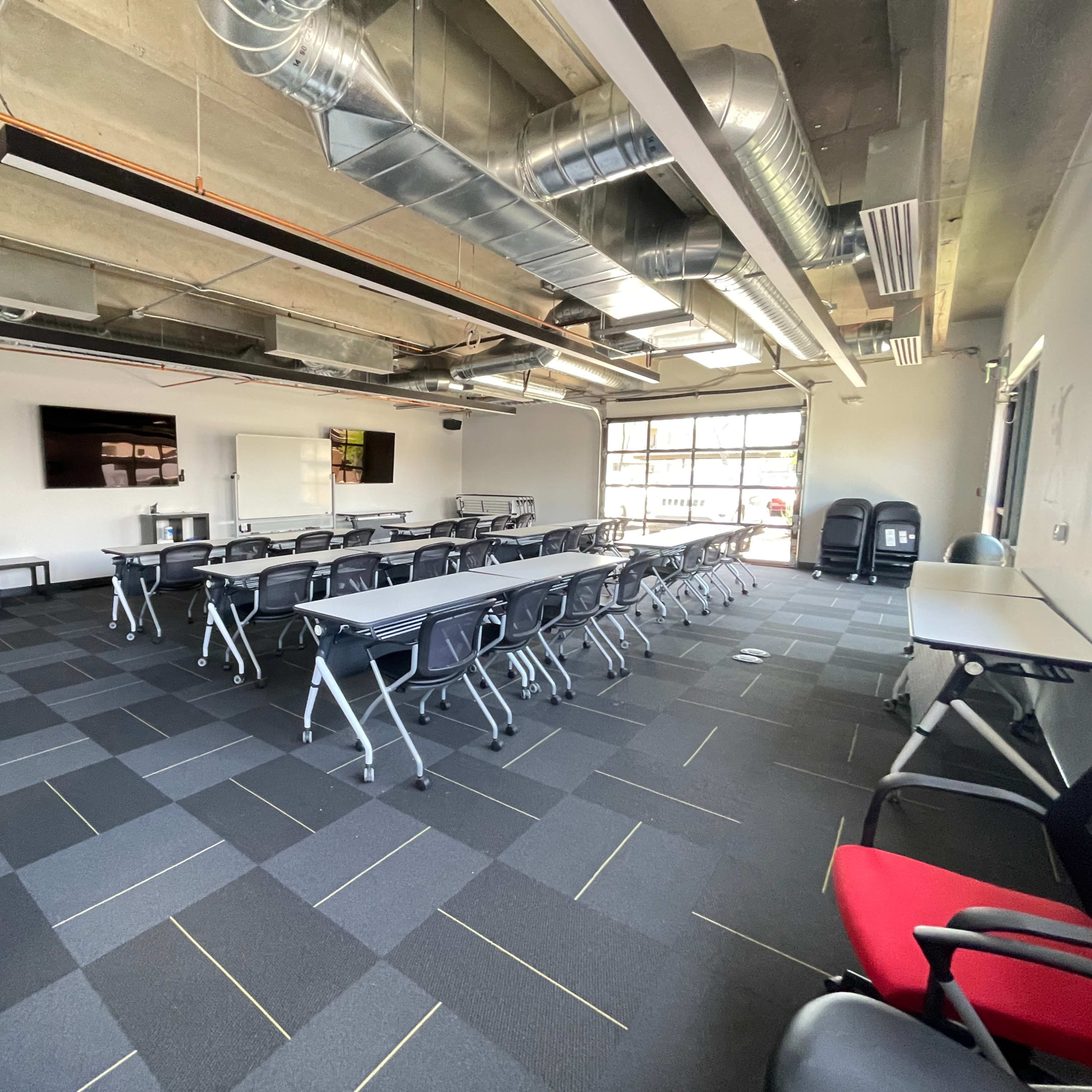A spacious classroom with rows of tables and chairs, large windows, and a presentation screen on the wall.