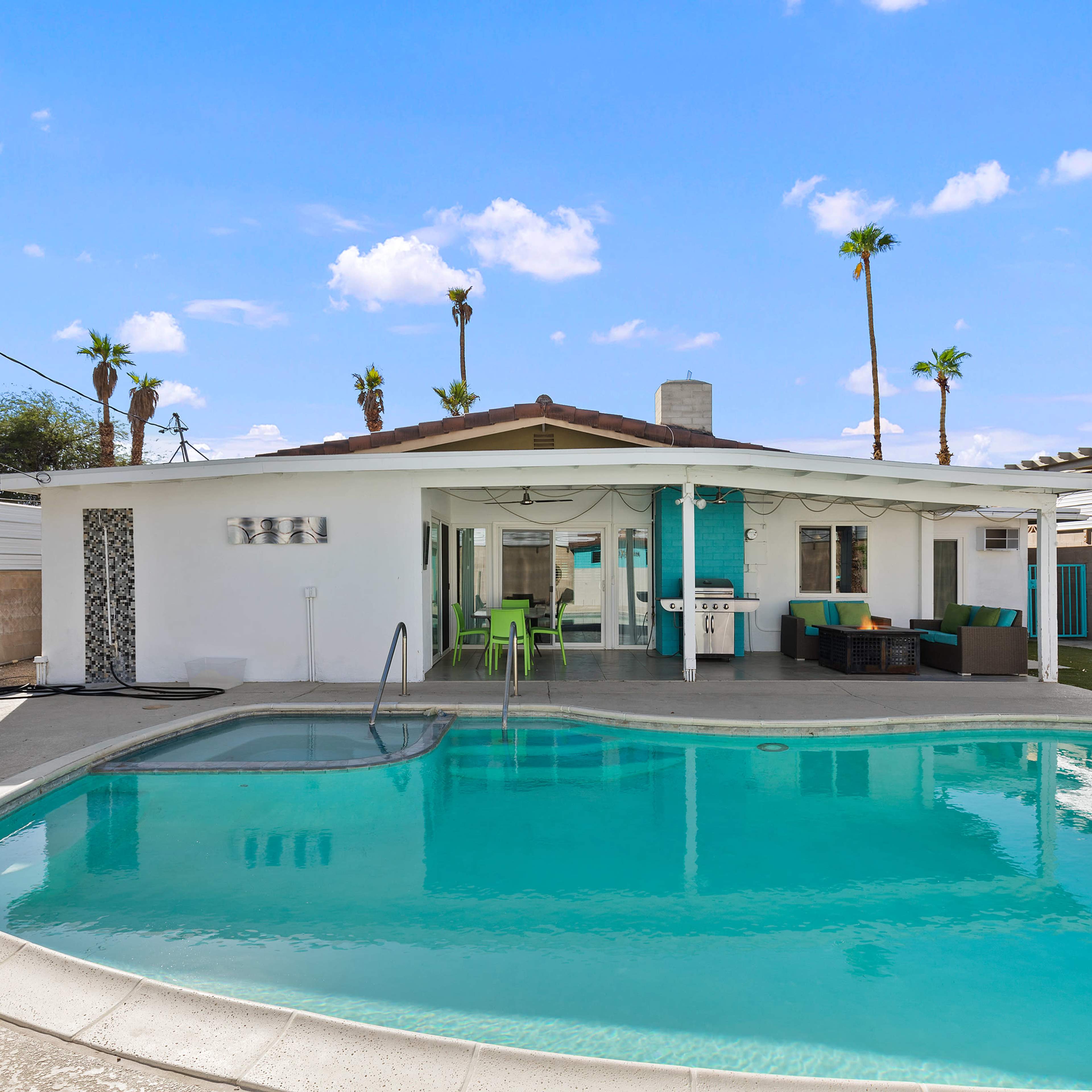 A residential backyard features a swimming pool next to a one-story house with palm trees in the background.