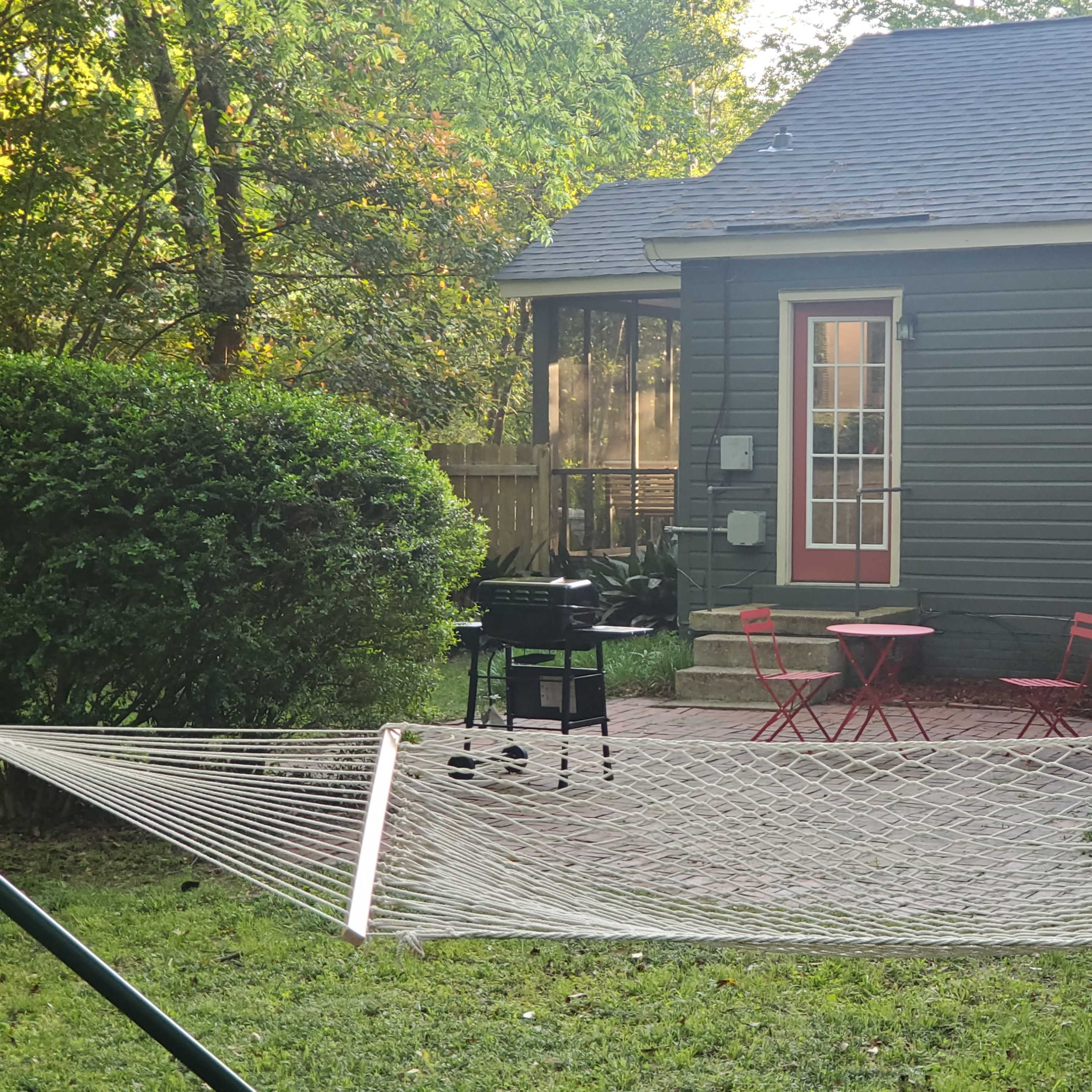 A hammock is strung between two supports in a yard with a grill and a small red table set in front of a green shrub and a gray house.