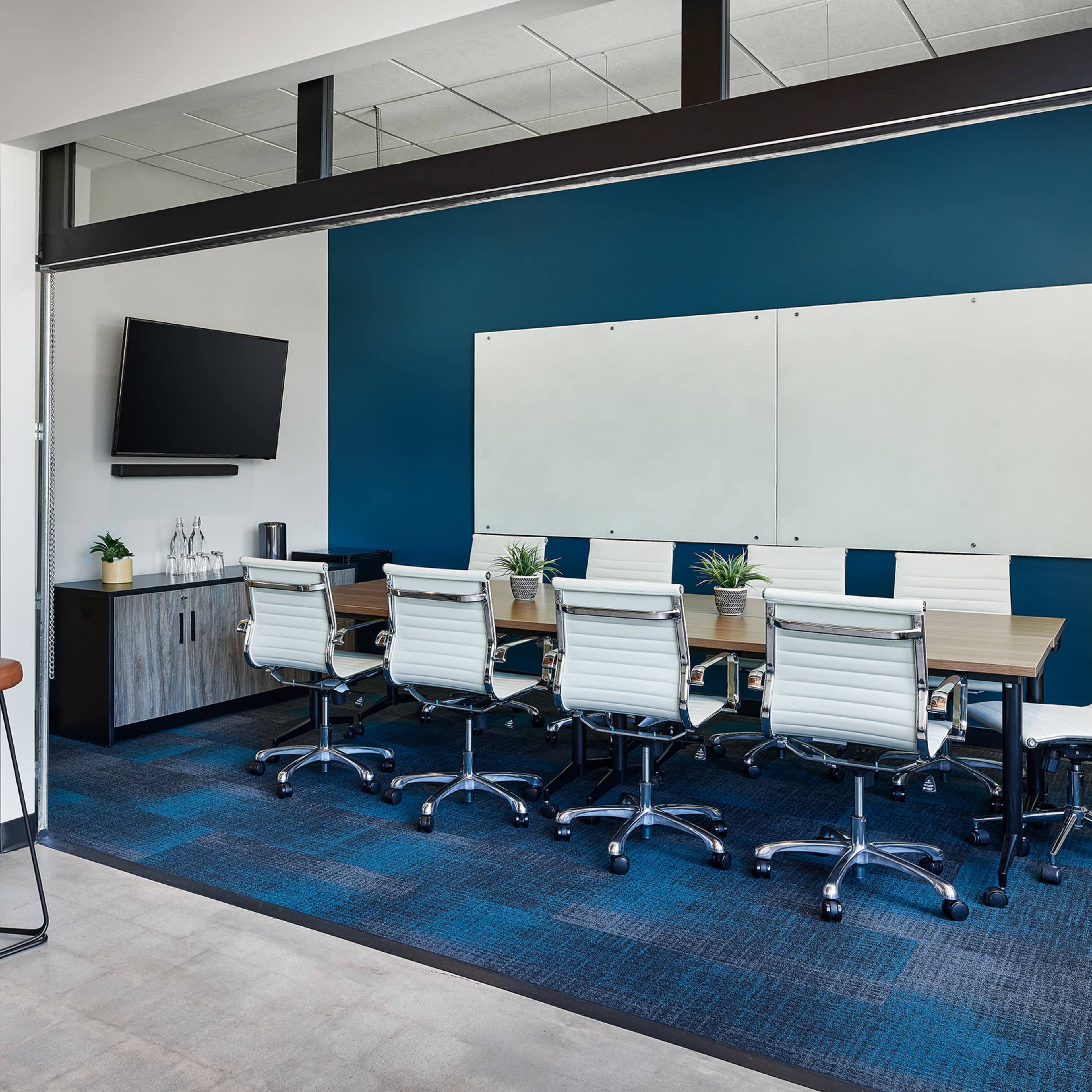 A modern conference room with a rectangular table surrounded by white swivel chairs, a television mounted on the wall, and a large whiteboard.