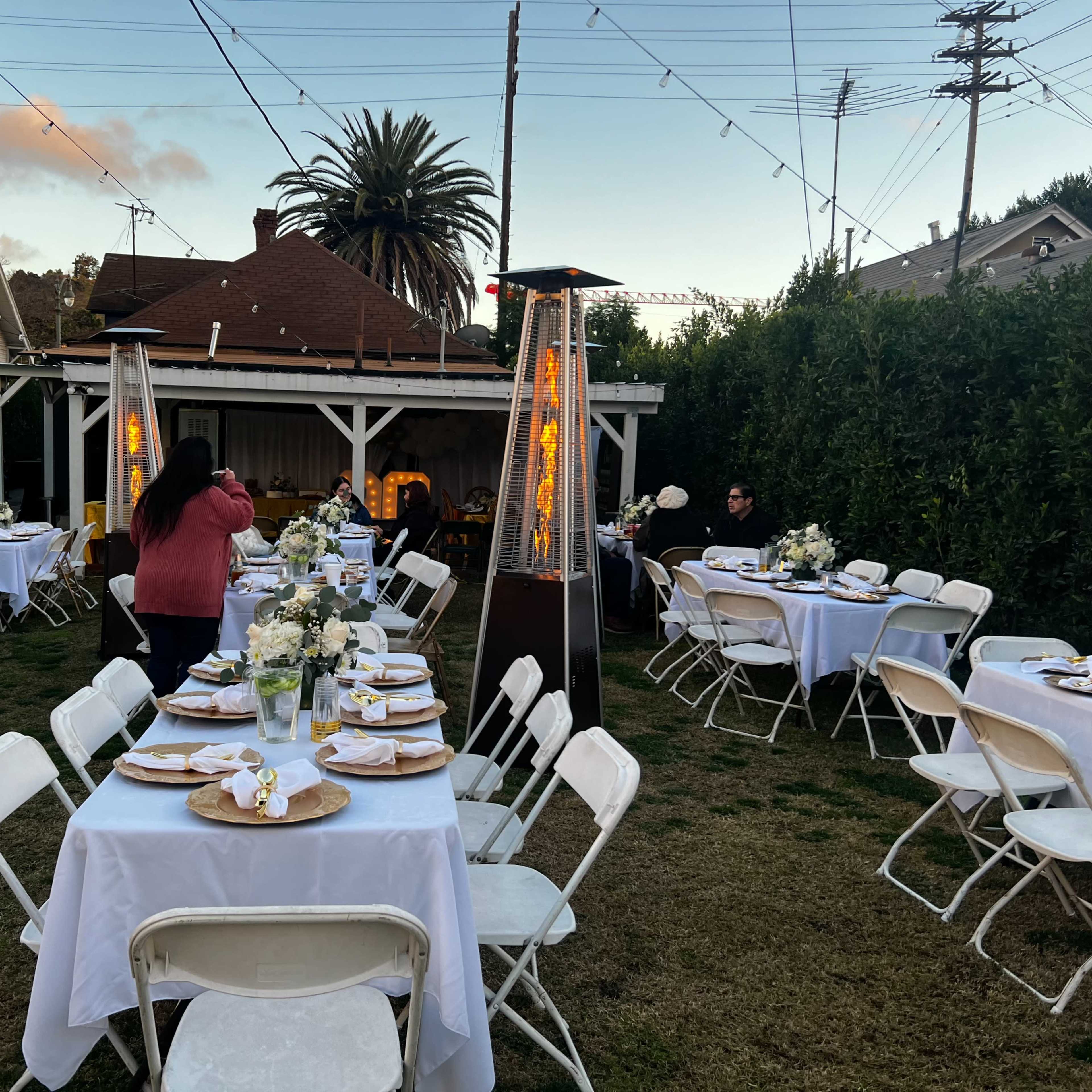 A backyard setting features several tables set with white tablecloths and plates, surrounded by string lights and greenery.