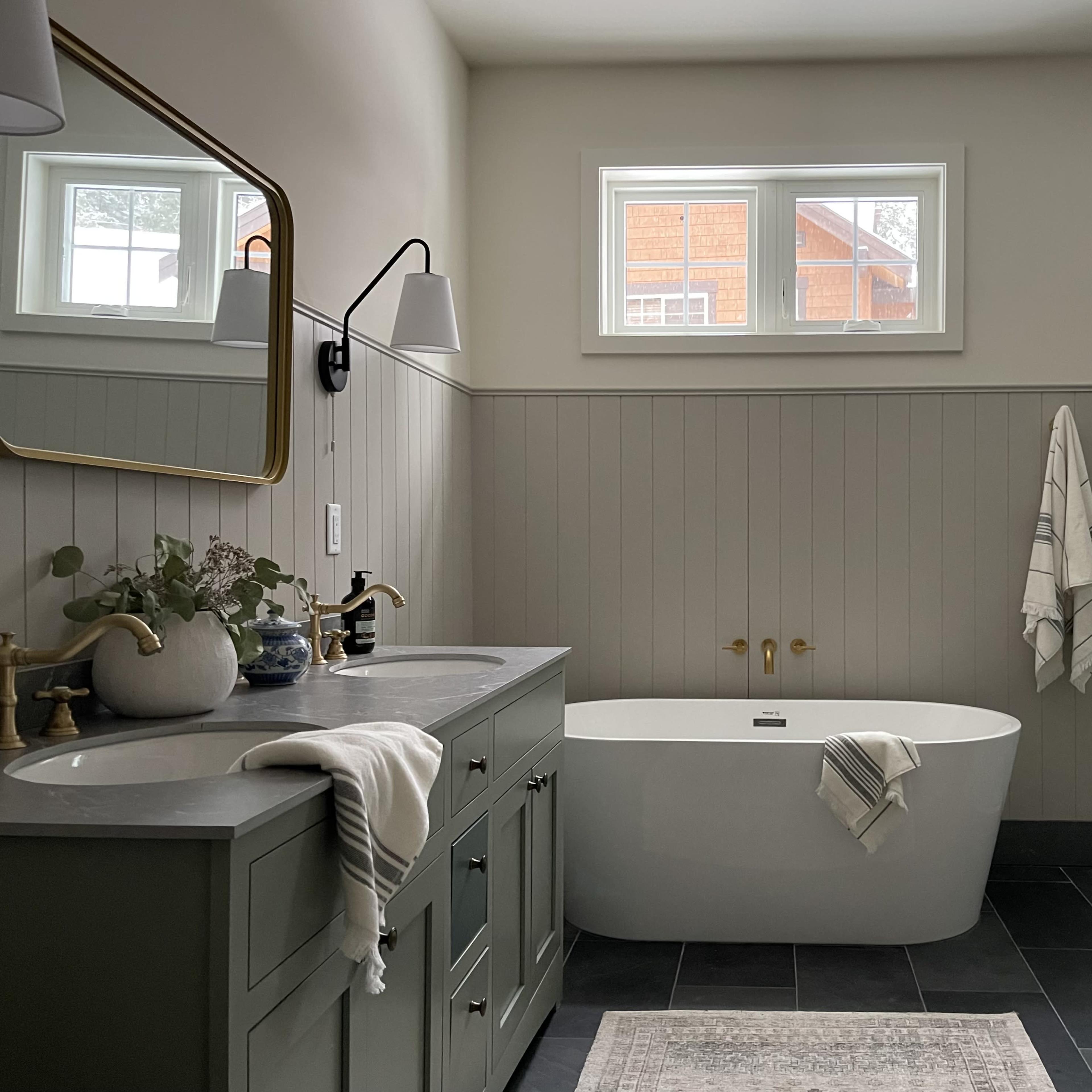 A modern bathroom features a freestanding bathtub, a glass shower stall, a double sink vanity with green cabinets, and large windows allowing natural light.