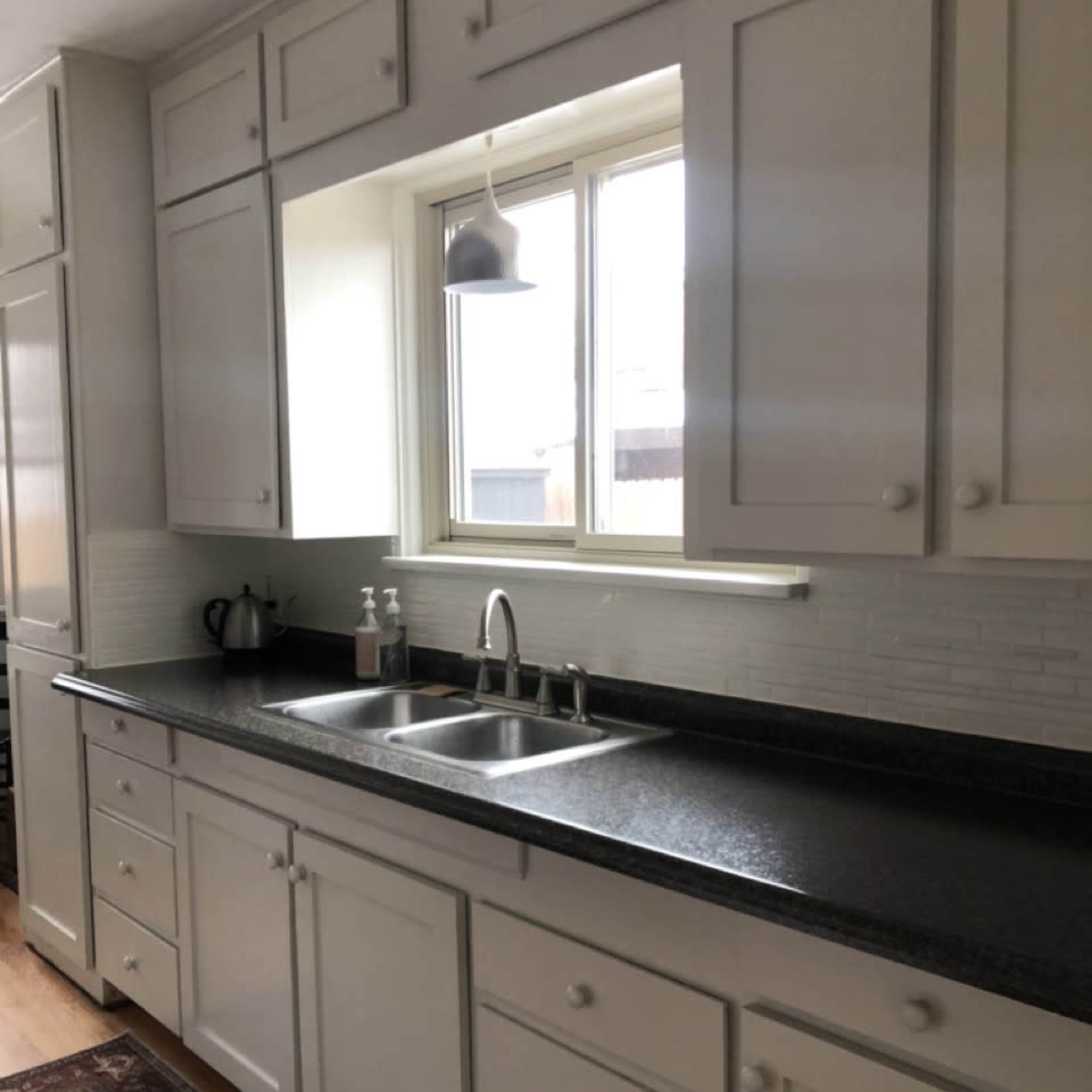 A kitchen with a gray countertop, double sink, and white cabinetry, featuring a window above the sink and a rug on the floor.
