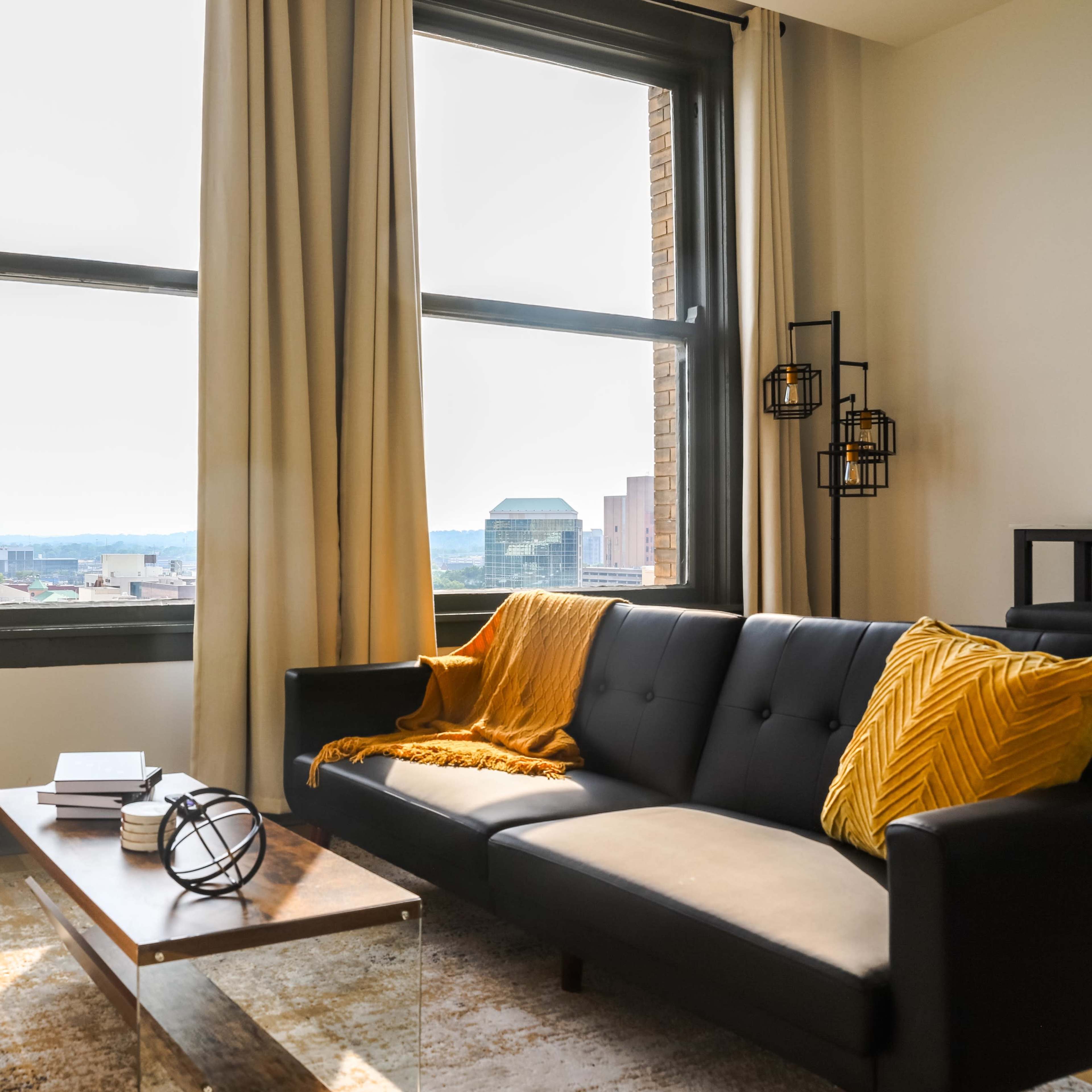 A modern living room features a black sofa with yellow accents, a wooden coffee table, and large windows allowing natural light to filter in, overlooking a cityscape.