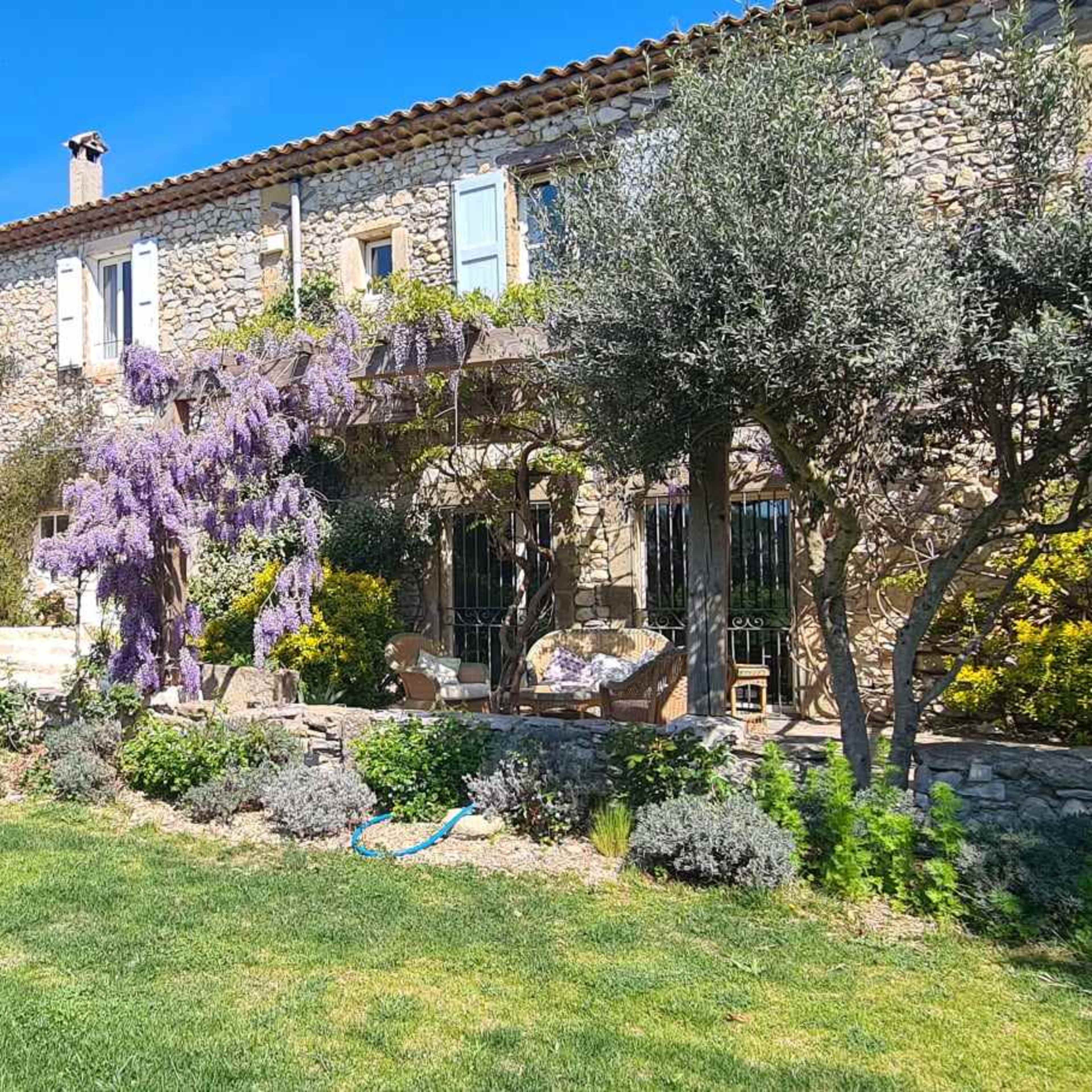 A stone house with colorful flowering plants and a lush green lawn is set against a clear blue sky.
