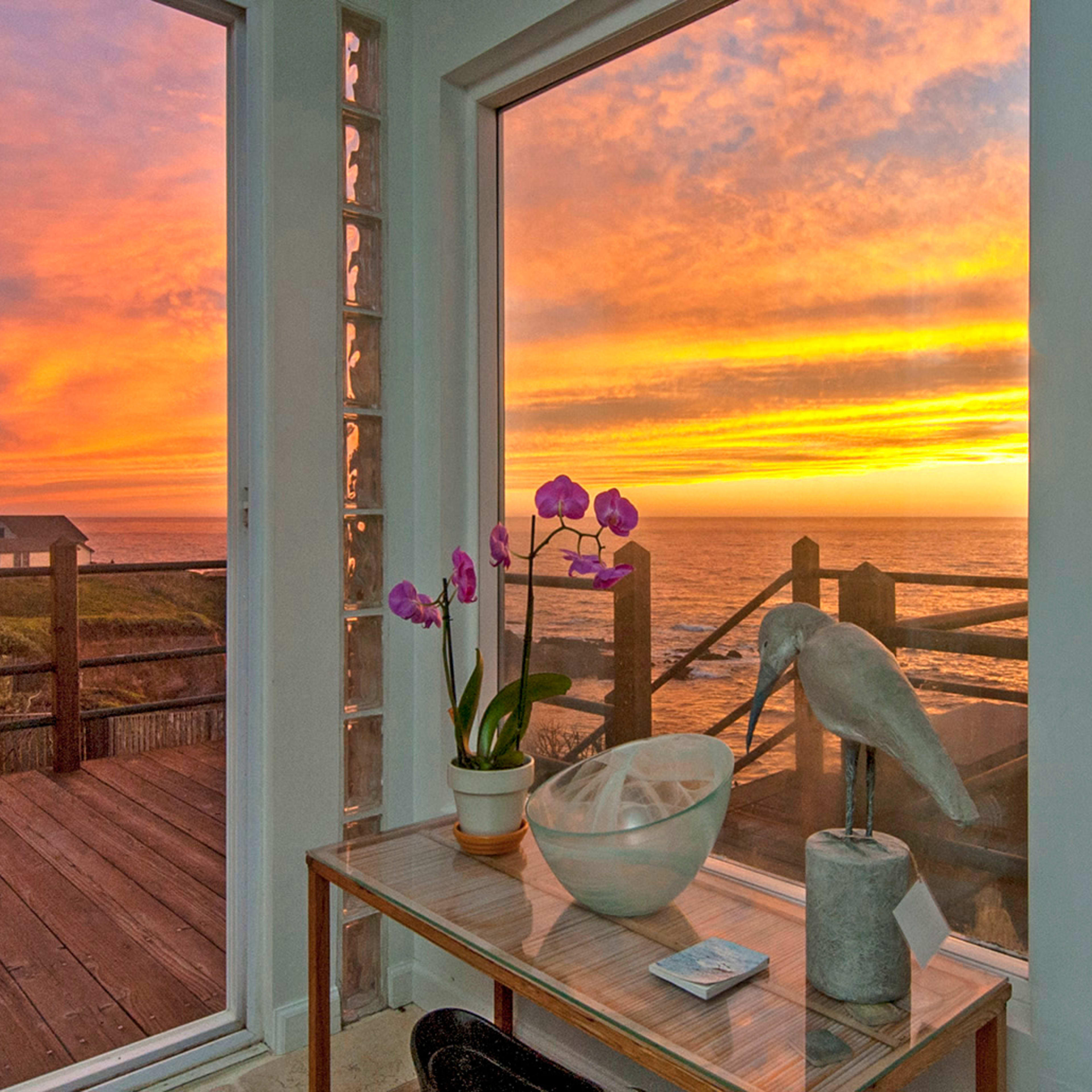 The scene features a coastal view at sunset through large windows, with a table displaying a vase of orchids and a decorative bird statue.