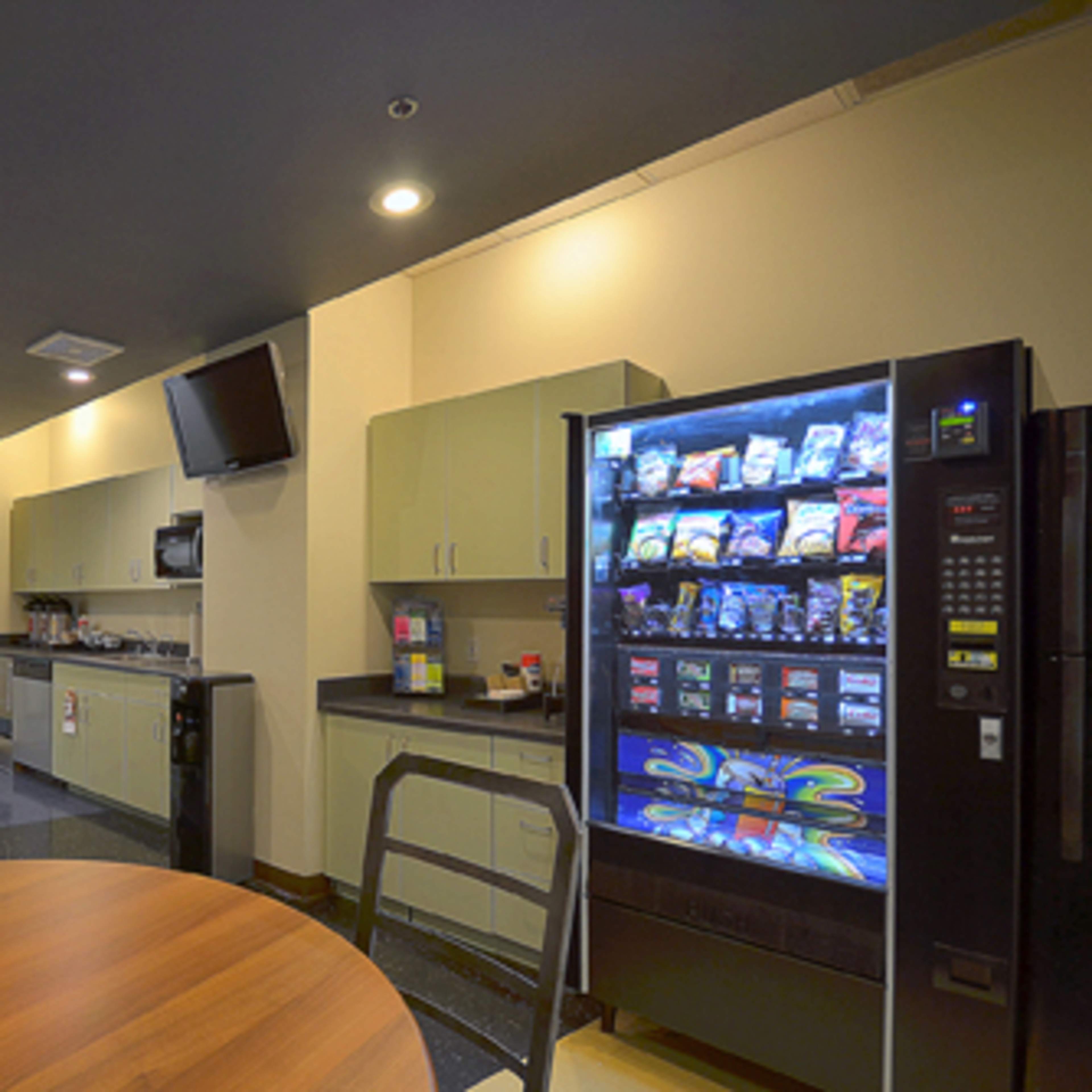 A break room with a vending machine, tables, and kitchen cabinets along two walls.