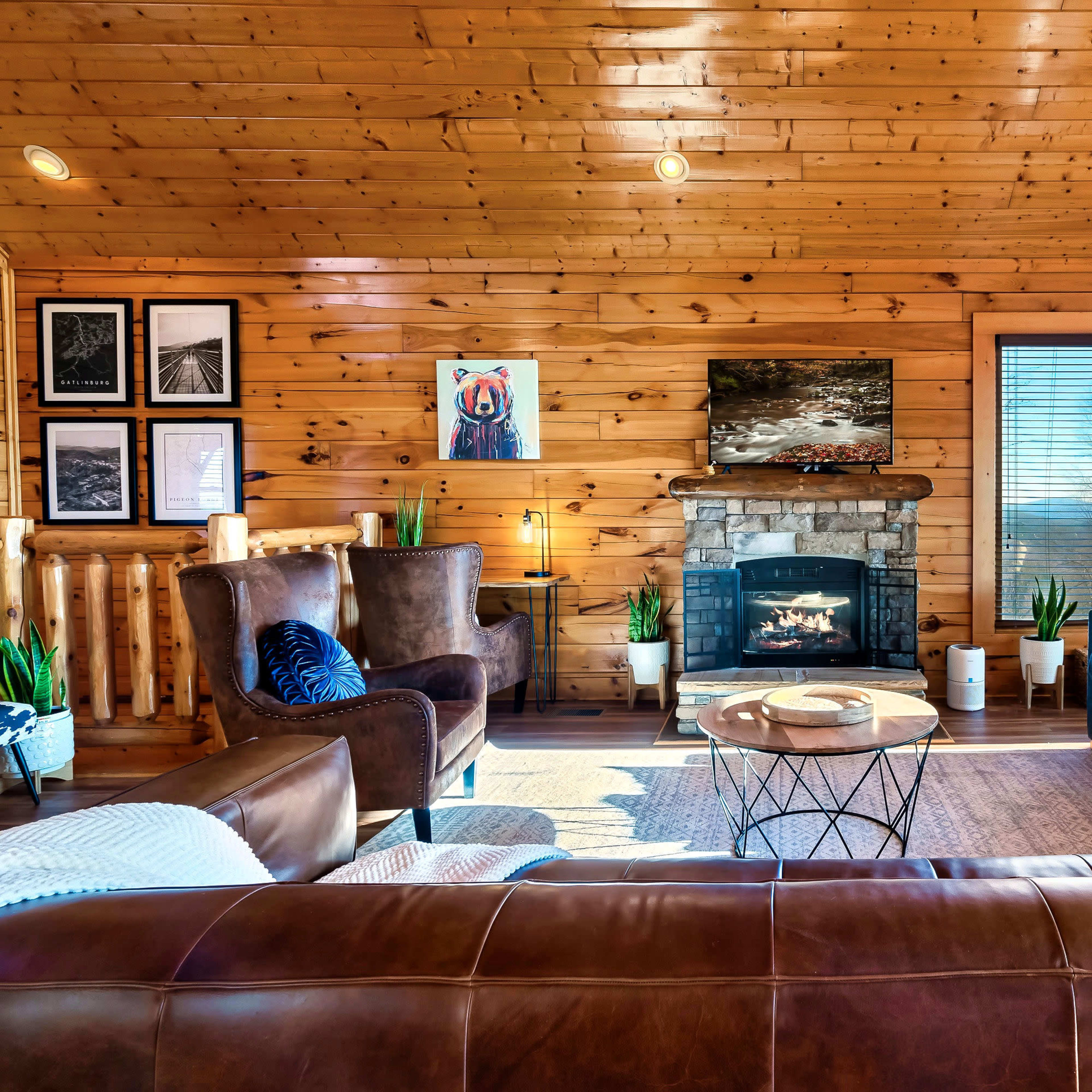 A warm and inviting wooden cabin interior with a stone fireplace, comfortable seating, and large windows revealing a scenic view.