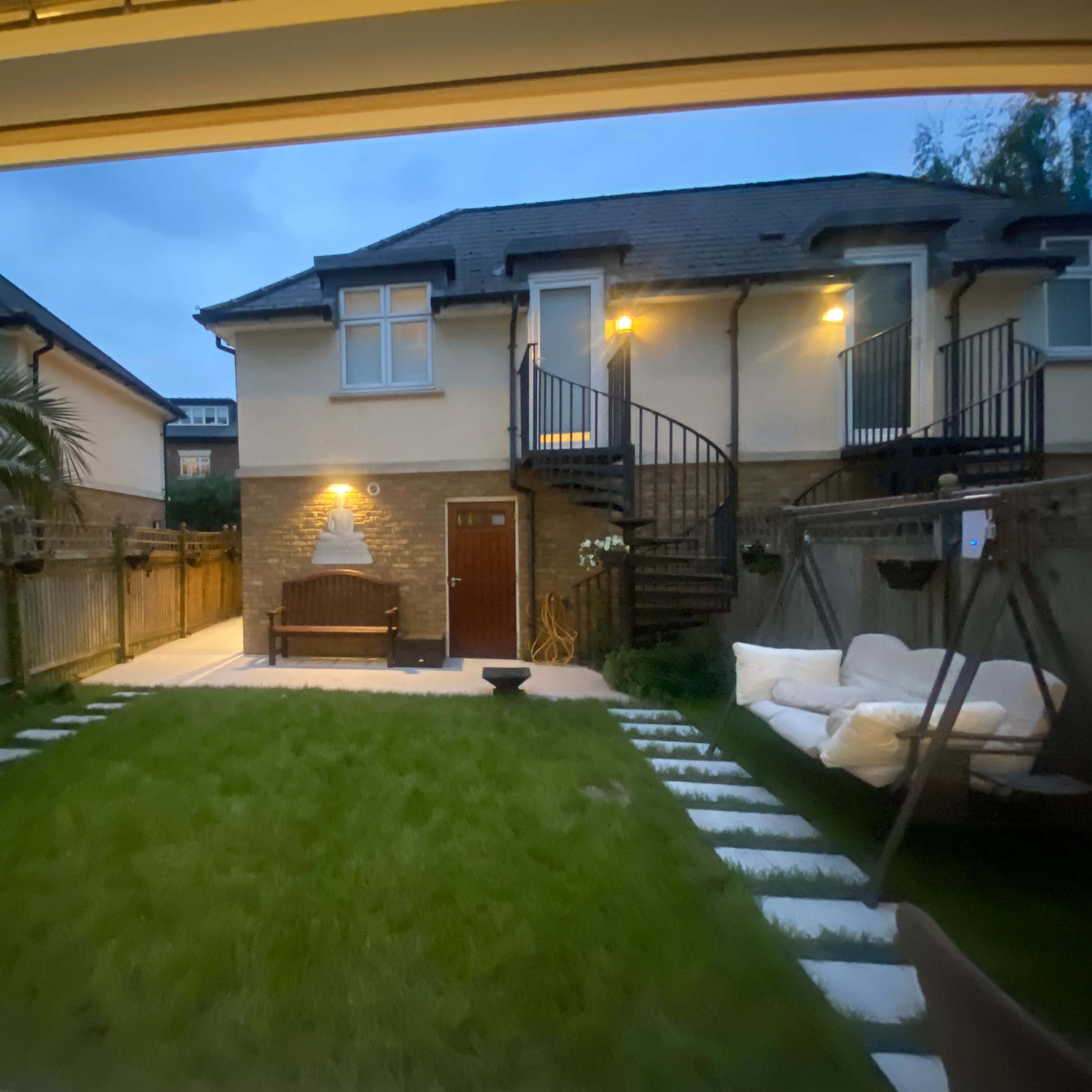 A well-maintained backyard features a grassy area with stone pathing, a bench, and a swing chair, bordered by two buildings with spiral staircases.