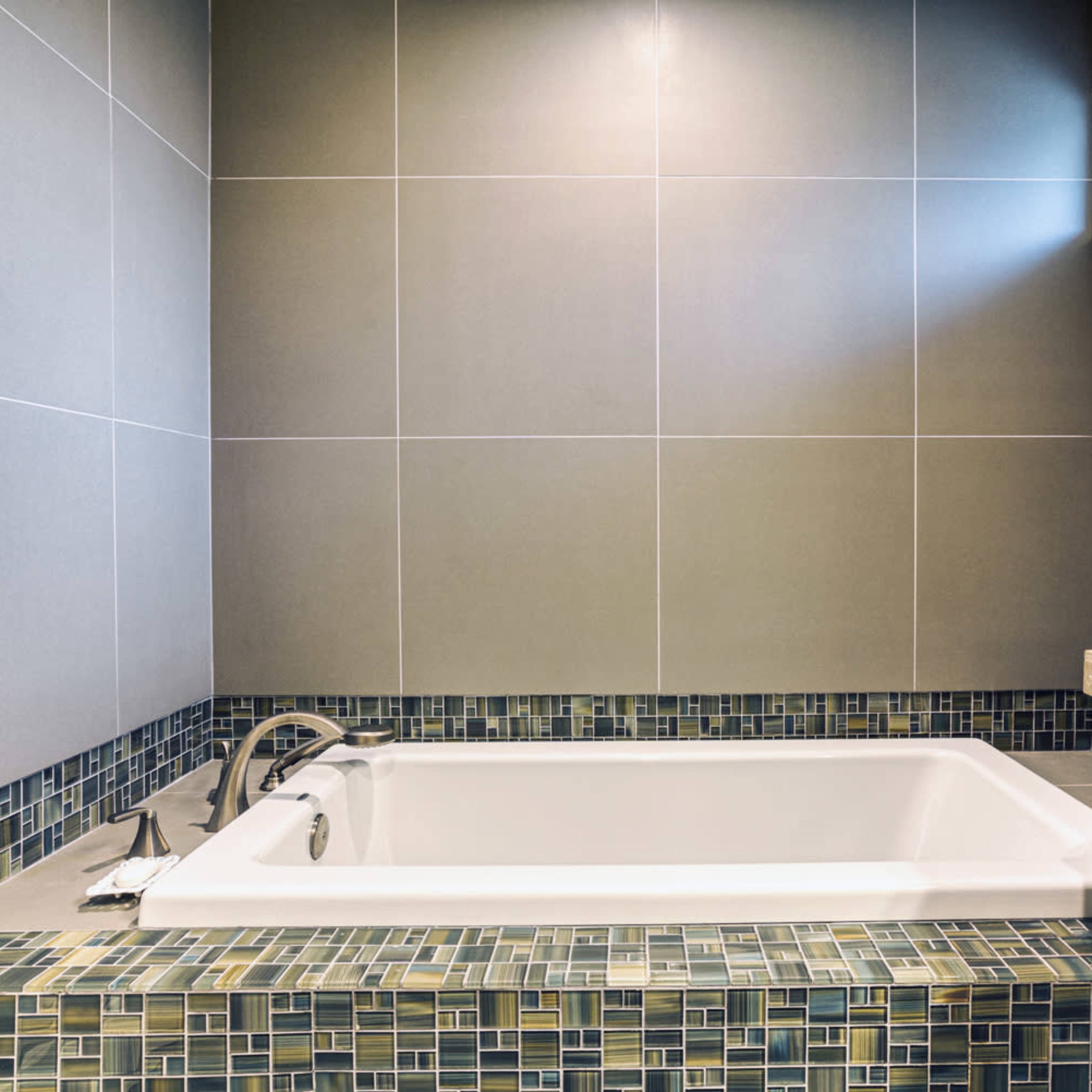 A modern bathtub surrounded by gray tiled walls and a mosaic tiled border.