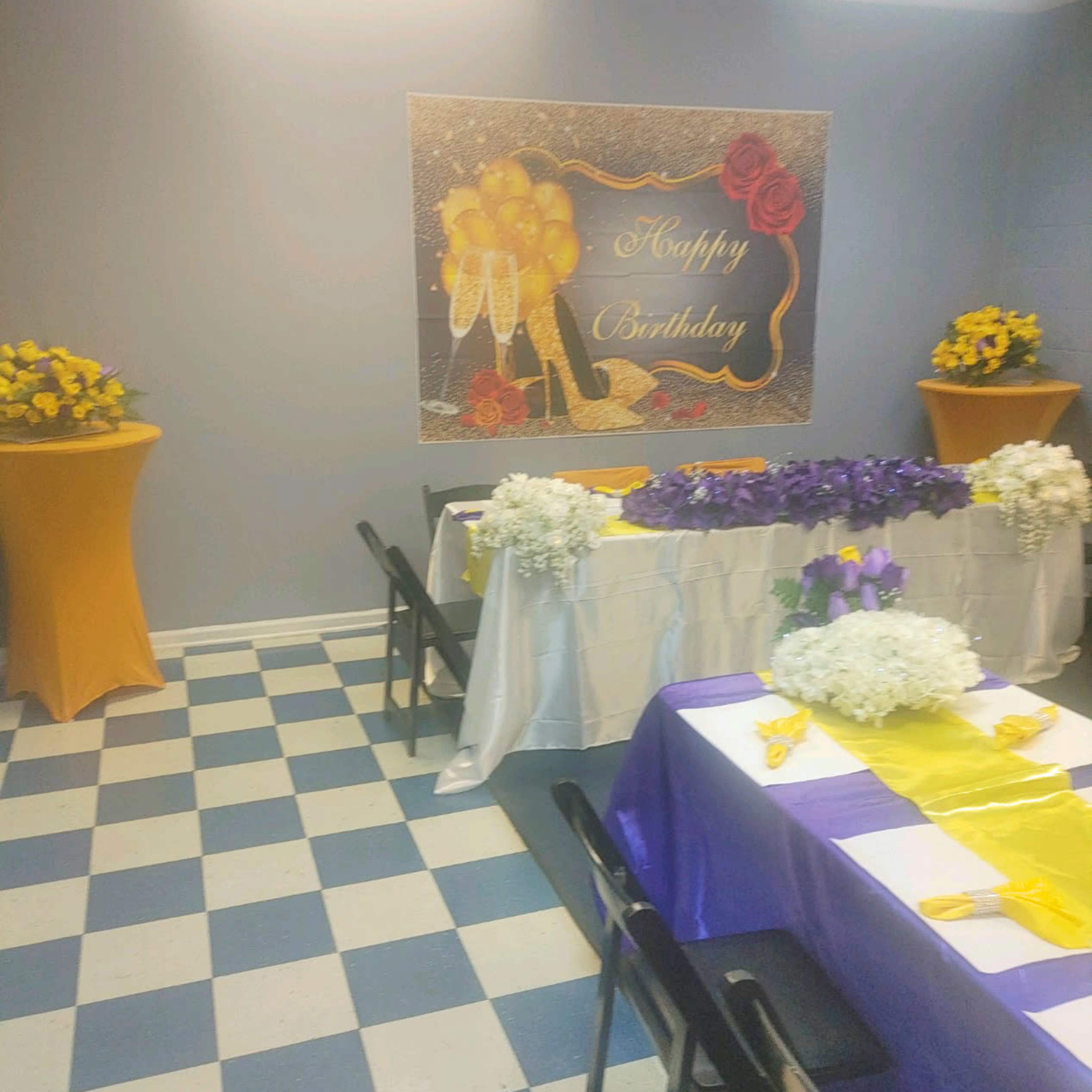 A decorated birthday celebration space features tables adorned with purple and yellow decorations, along with floral arrangements and a backdrop that reads "Happy Birthday."