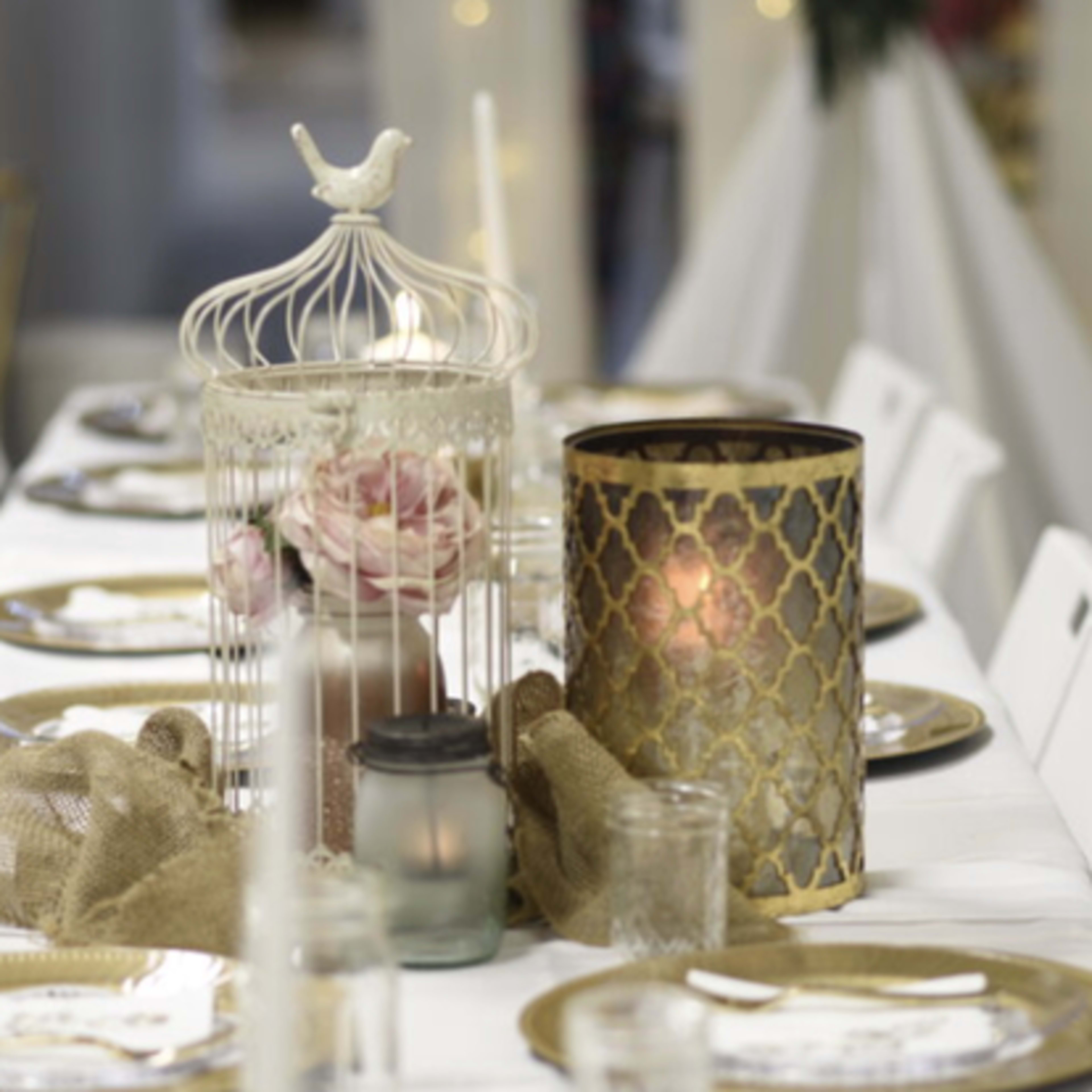 A long dining table is elegantly set with gold-rimmed plates, decorative candles, and a decorative birdcage centerpiece.