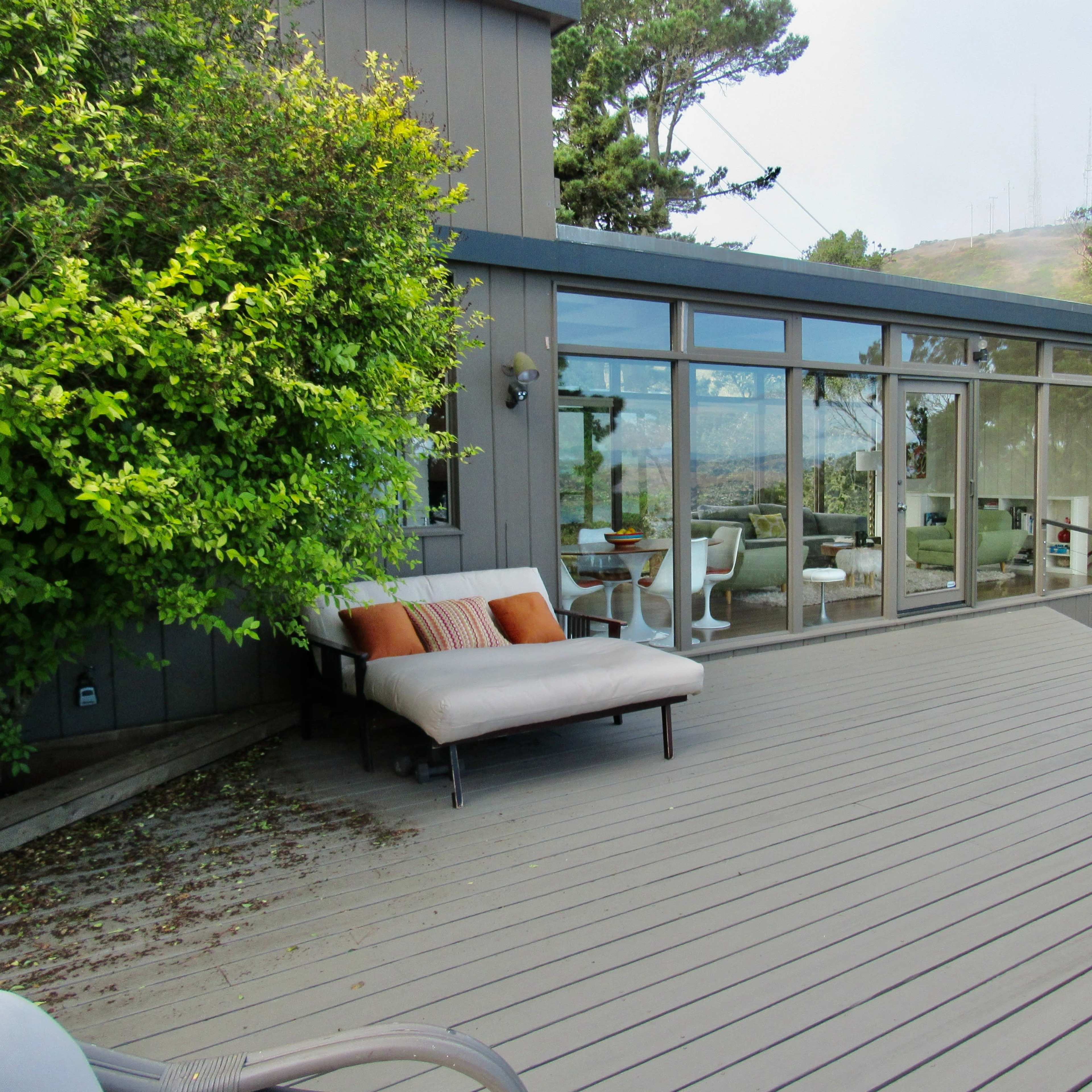 A modern deck features a lounging area with a couch and a view of large glass windows reflecting a hillside.