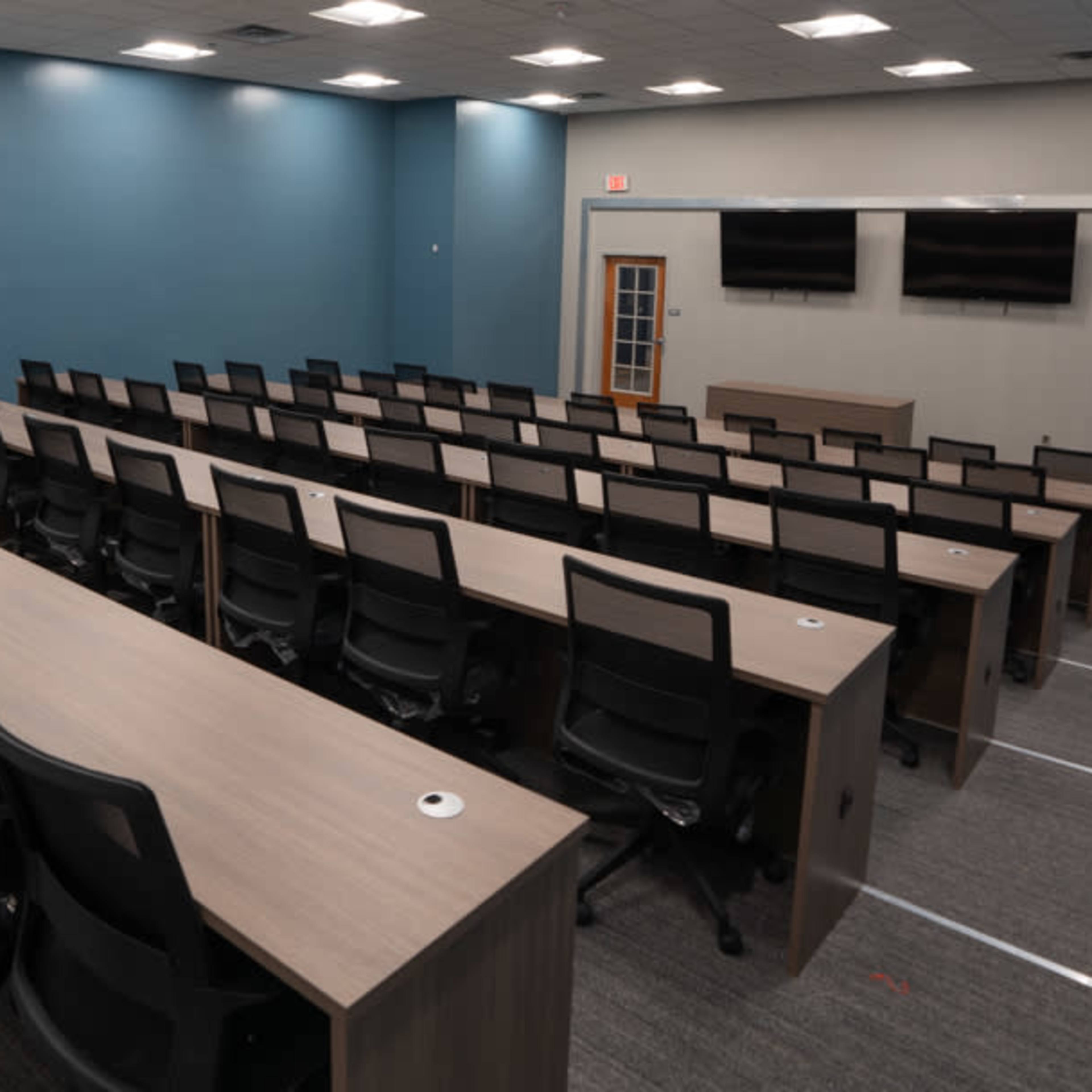 A modern classroom or conference room with rows of long tables and chairs arranged facing a presentation area, featuring two wall-mounted screens.