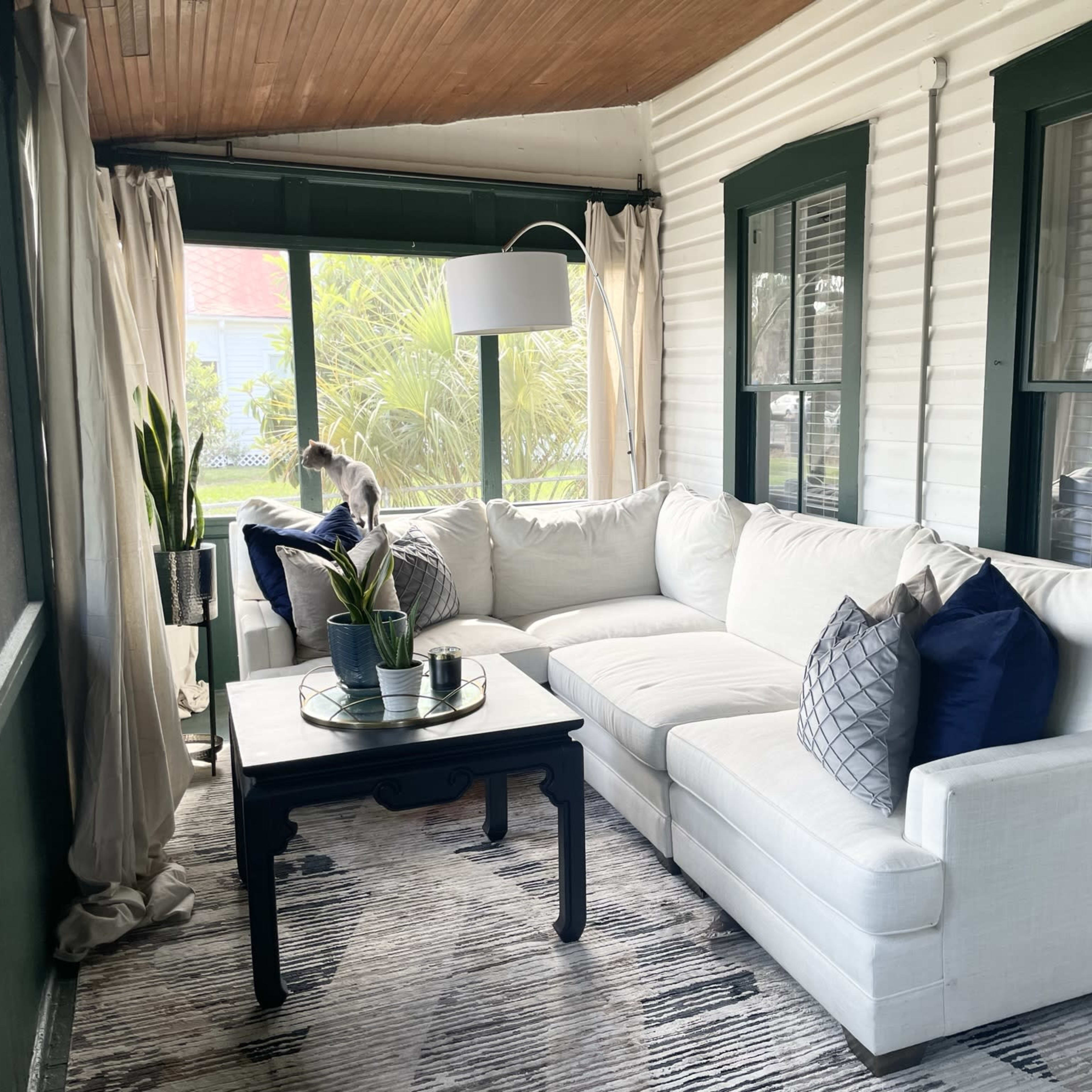 A sunlit screened porch featuring a white sectional sofa with navy and gray pillows, a dark coffee table, and potted plants, surrounded by windows and light-colored curtains.