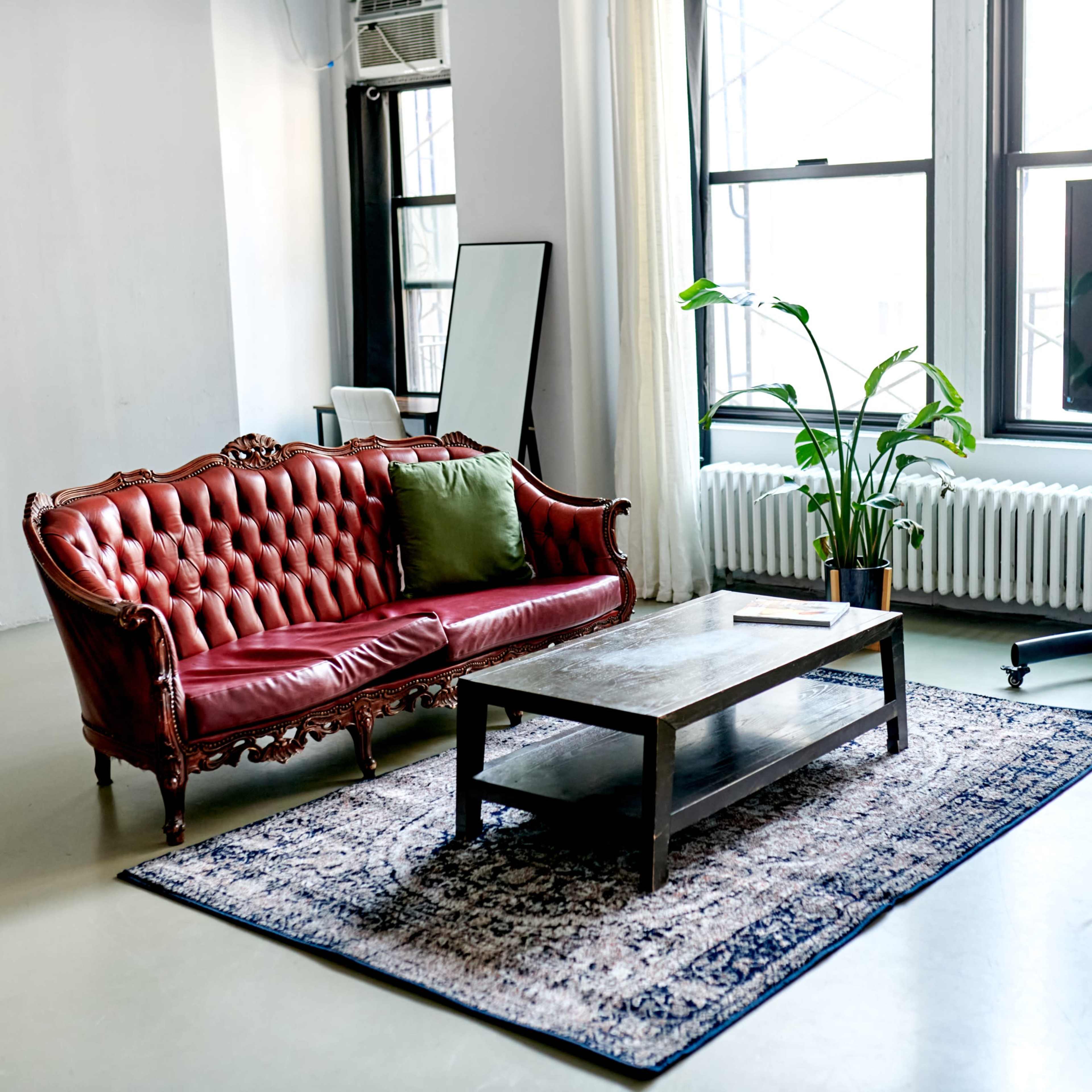 A living room setting with a red tufted sofa, a wooden coffee table on a patterned rug, and a large television on a mobile stand beside a potted plant near a window.