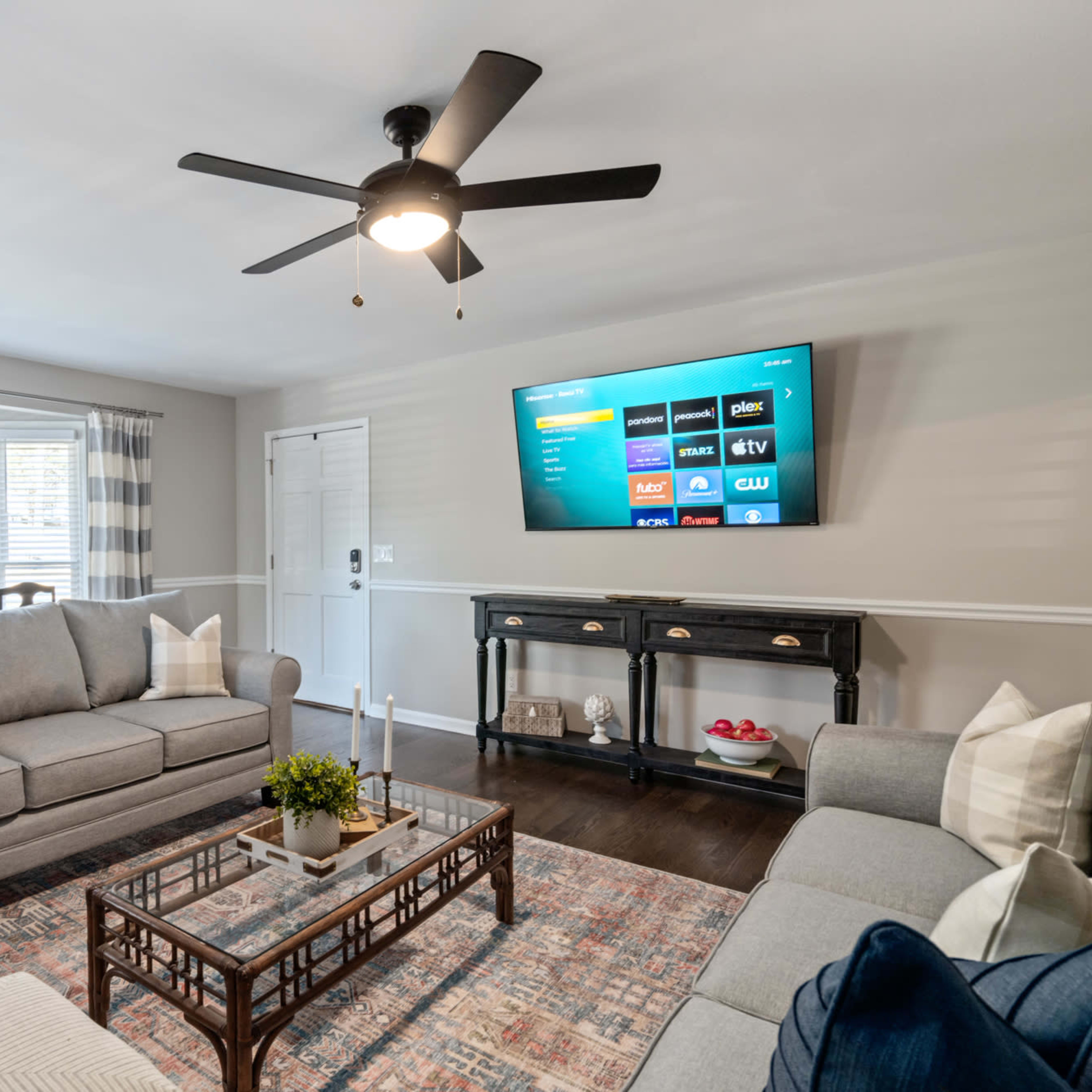 A well-furnished living room features a gray sofa set, a coffee table, a television mounted on the wall, and large windows dressed with curtains.