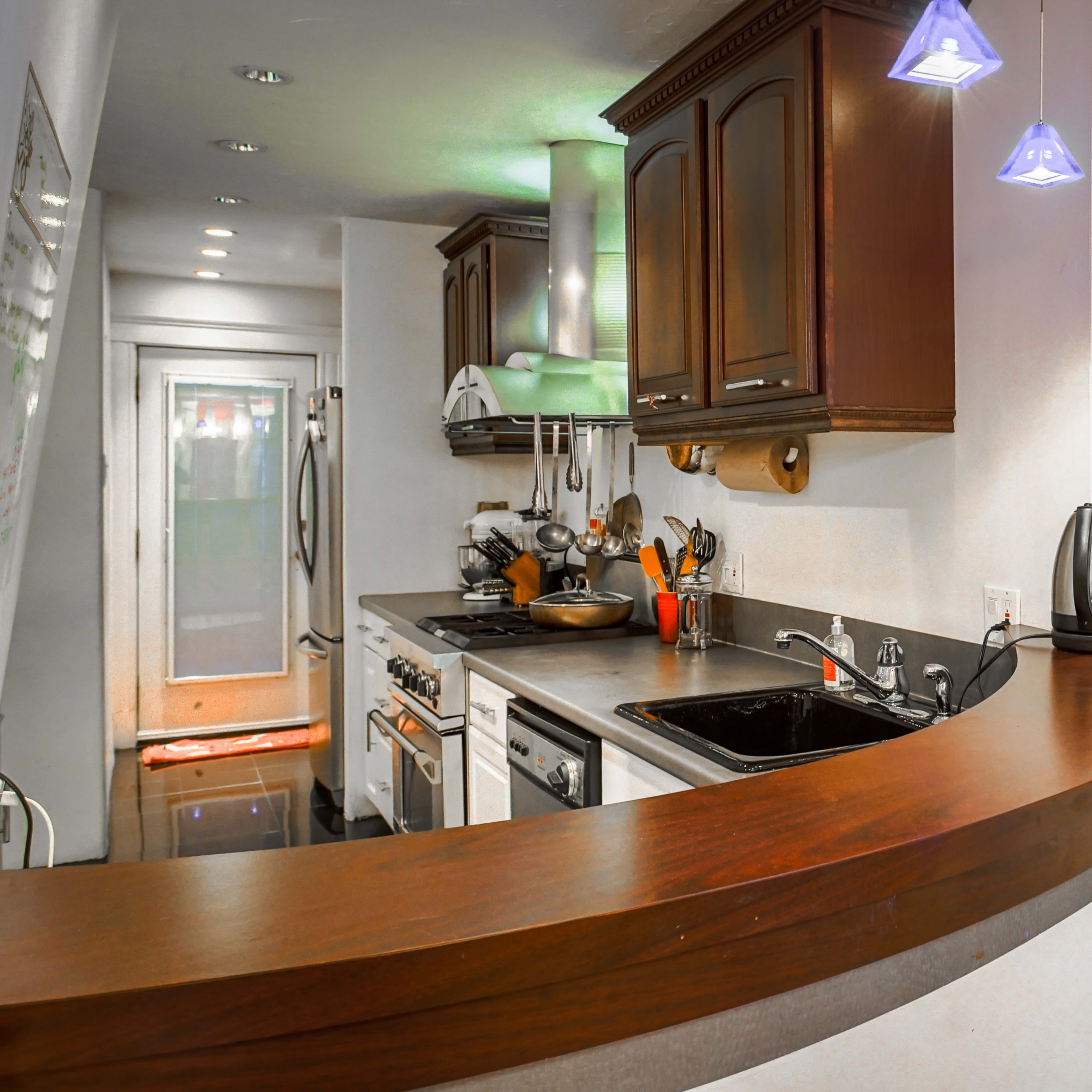 A kitchen with dark cabinetry, a curved wooden counter, and a hanging potted plant near a window, featuring kitchen appliances and utensils.