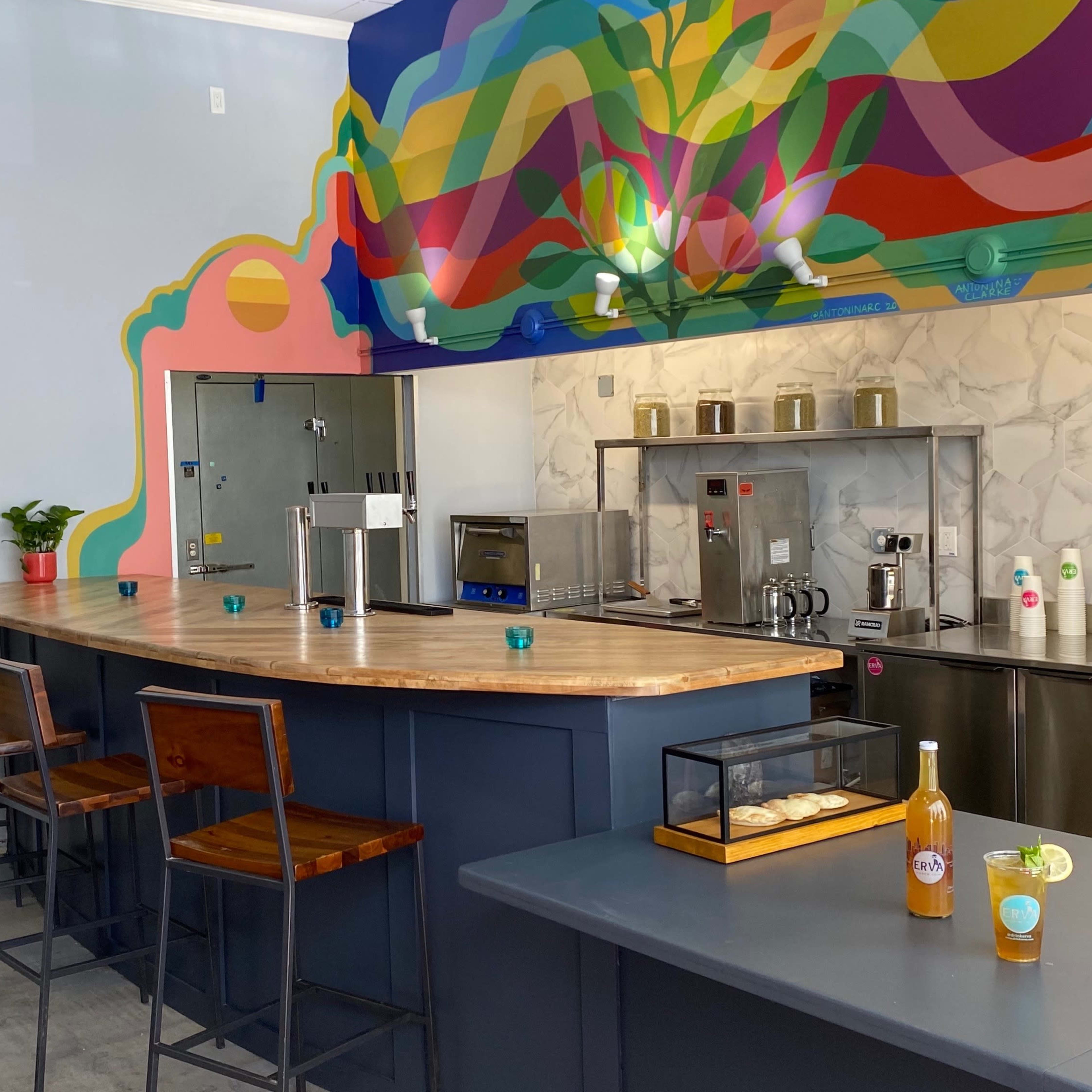 A modern cafe interior featuring a curved wooden bar with high stools, colorful mural artwork on the wall, and various kitchen appliances in a stainless steel setup.