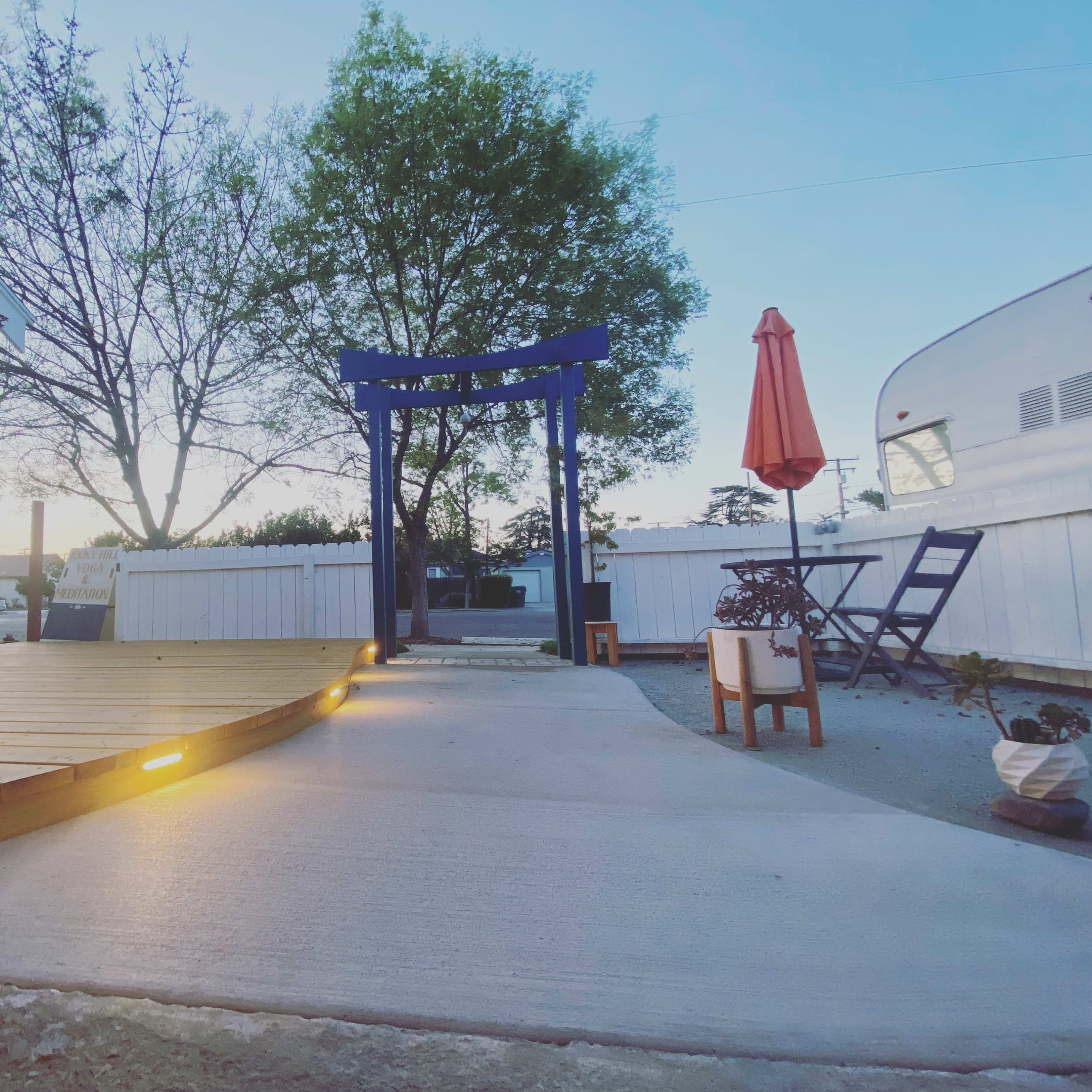 A backyard area featuring a wooden deck, a blue torii gate, an umbrella-shaded table, and a concrete pathway leading to the entrance.