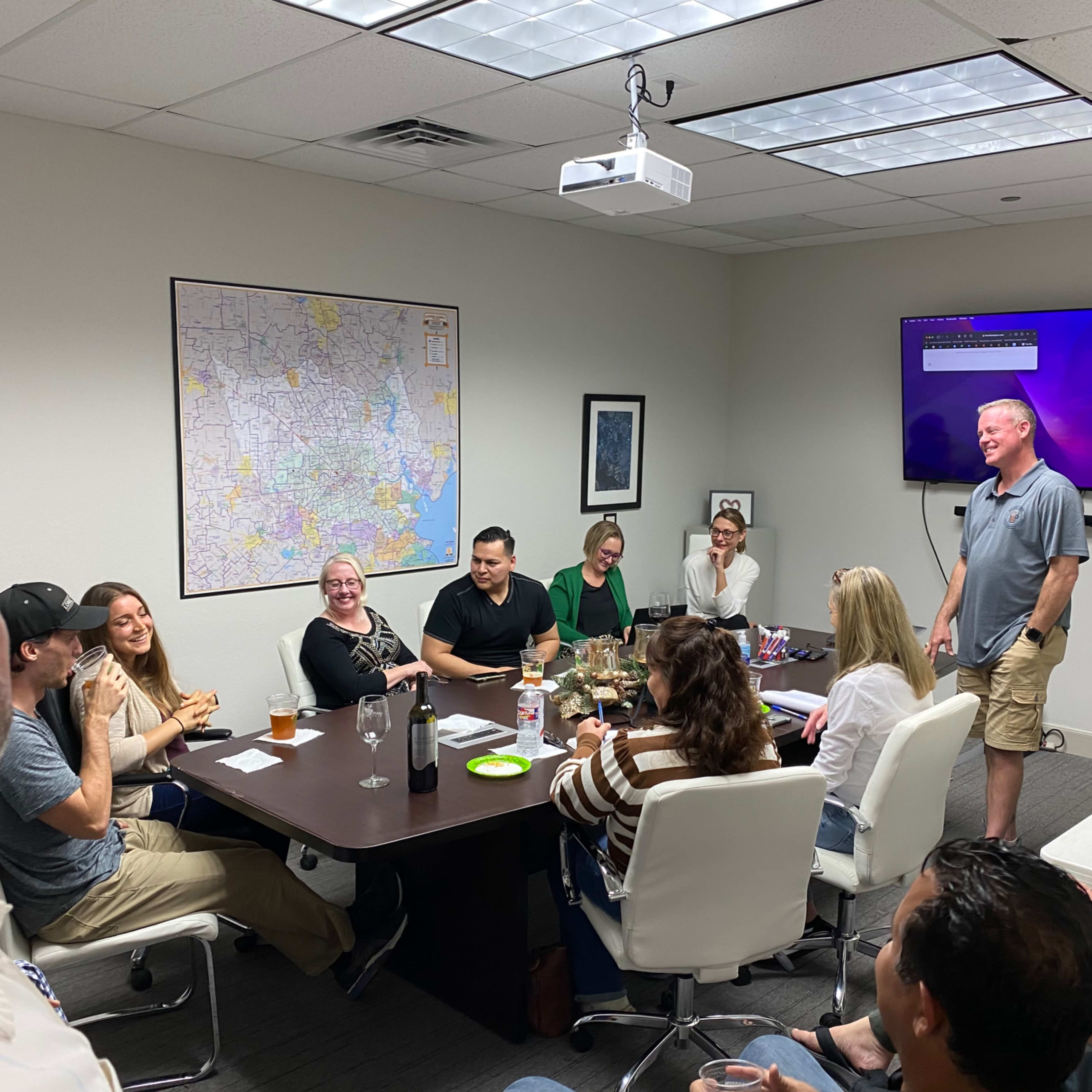 A group of people gathers around a conference table in a meeting room, engaging in conversation while a presentation screen displays information.