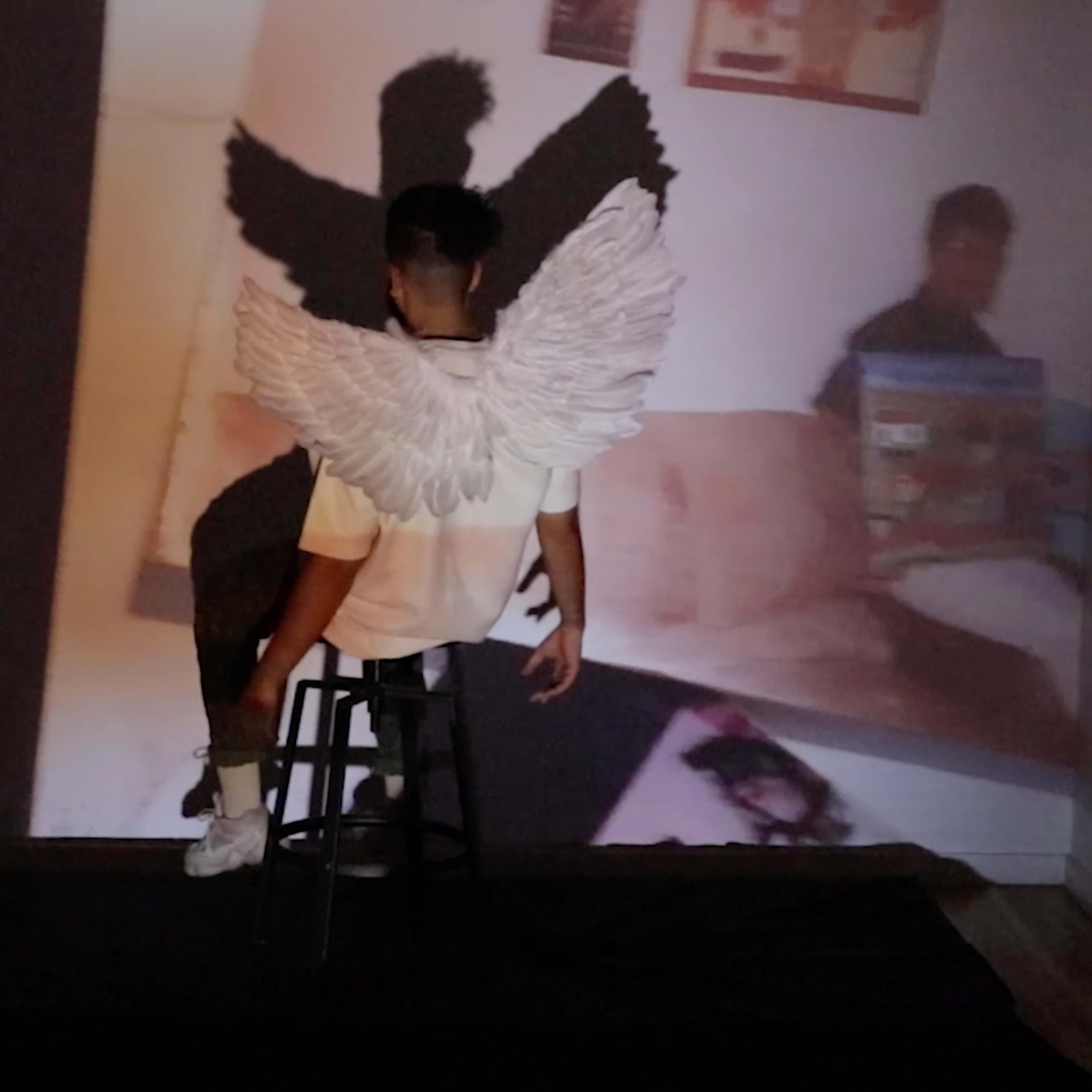 A person with large white wings sits on a stool in front of projected images on the wall.