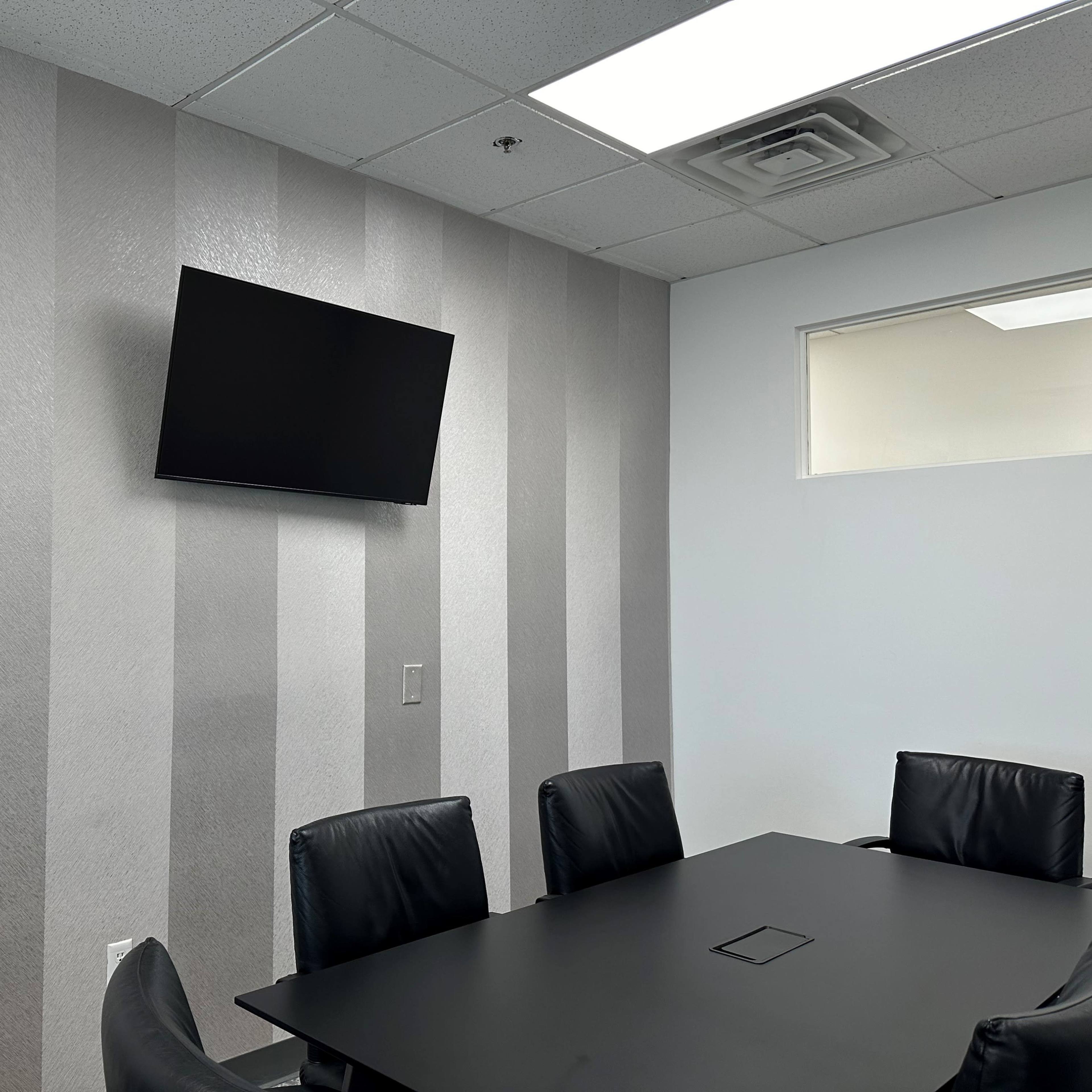 A conference room features a black television mounted on a striped gray wall and a rectangular table surrounded by black leather chairs.