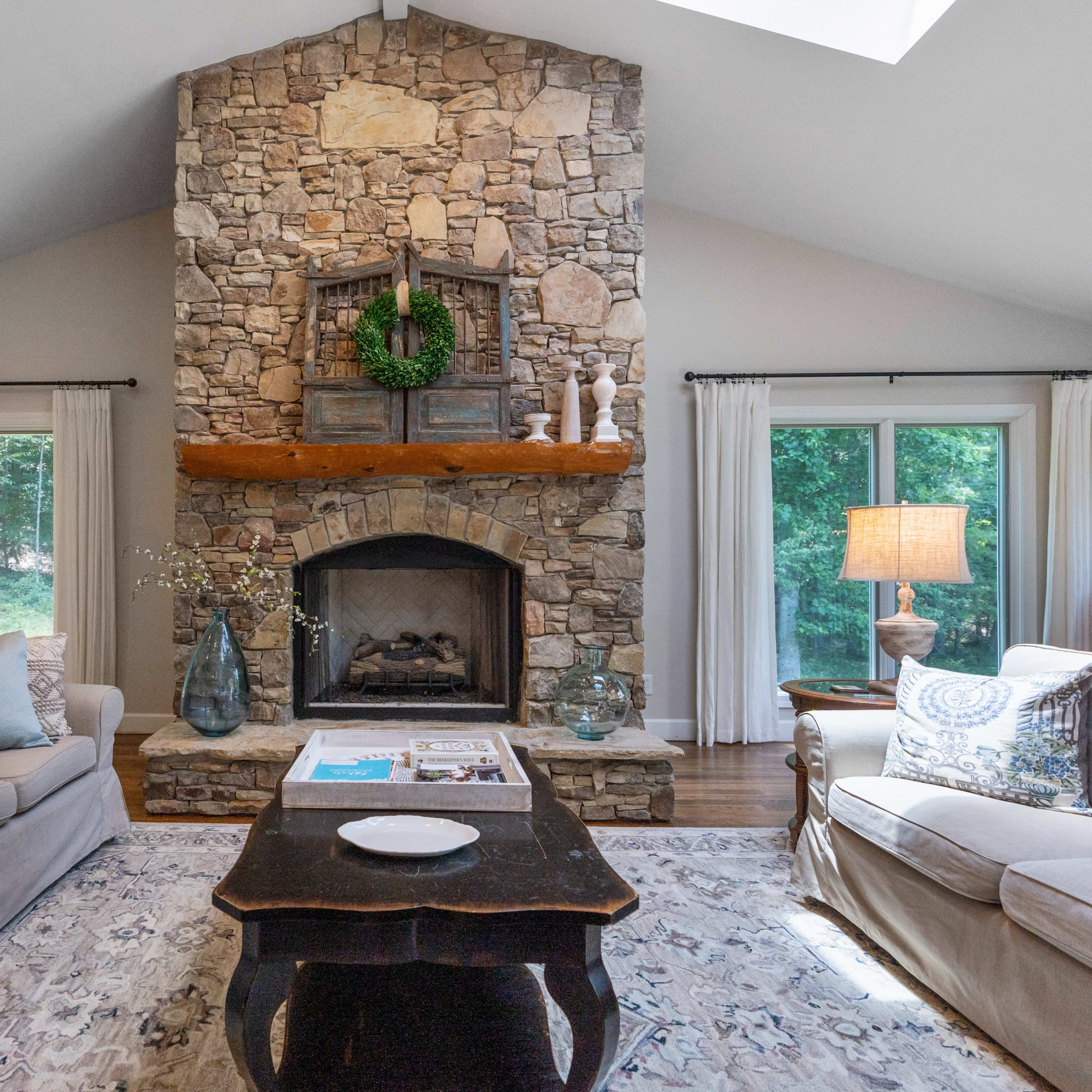 The living room features a large stone fireplace with a wooden mantel, surrounded by two sofas and natural light from large windows.