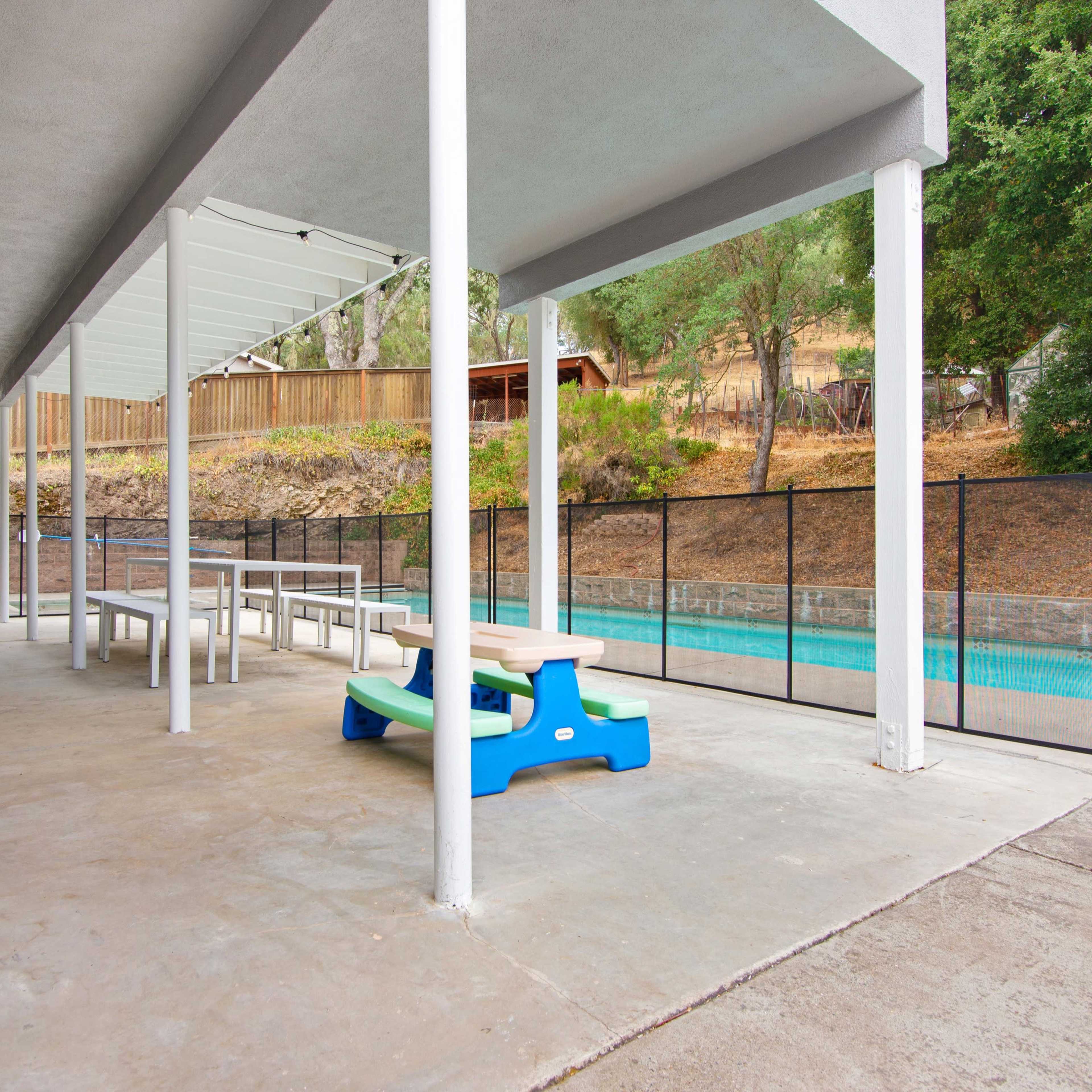 A patio area with a table and chairs, a children's play table, and a swimming pool enclosed by a fence.
