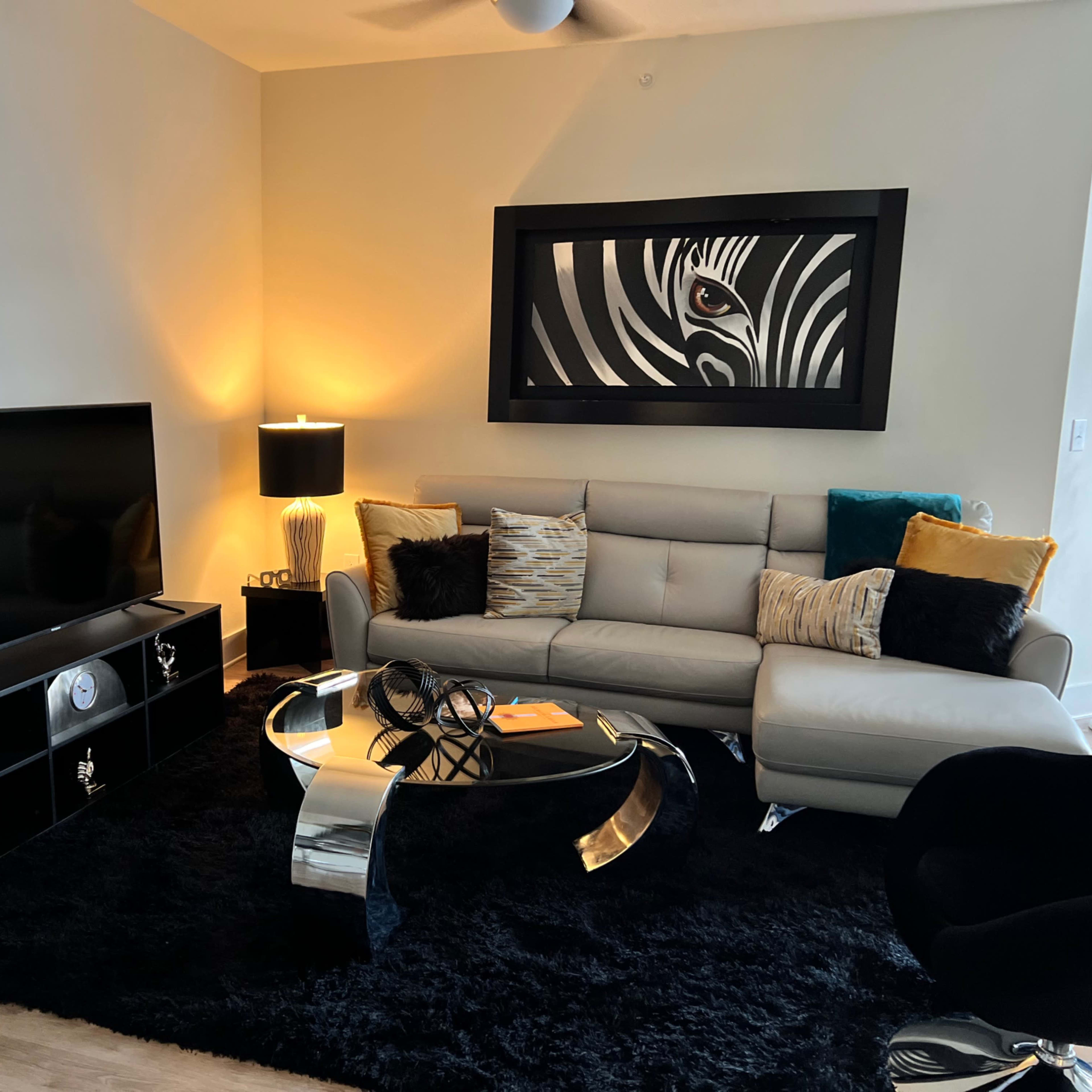 A modern living room featuring a light-colored sofa, a black entertainment unit with a large television, a glass coffee table, and decorative accents including a zebra-patterned artwork and a metallic sculpture.