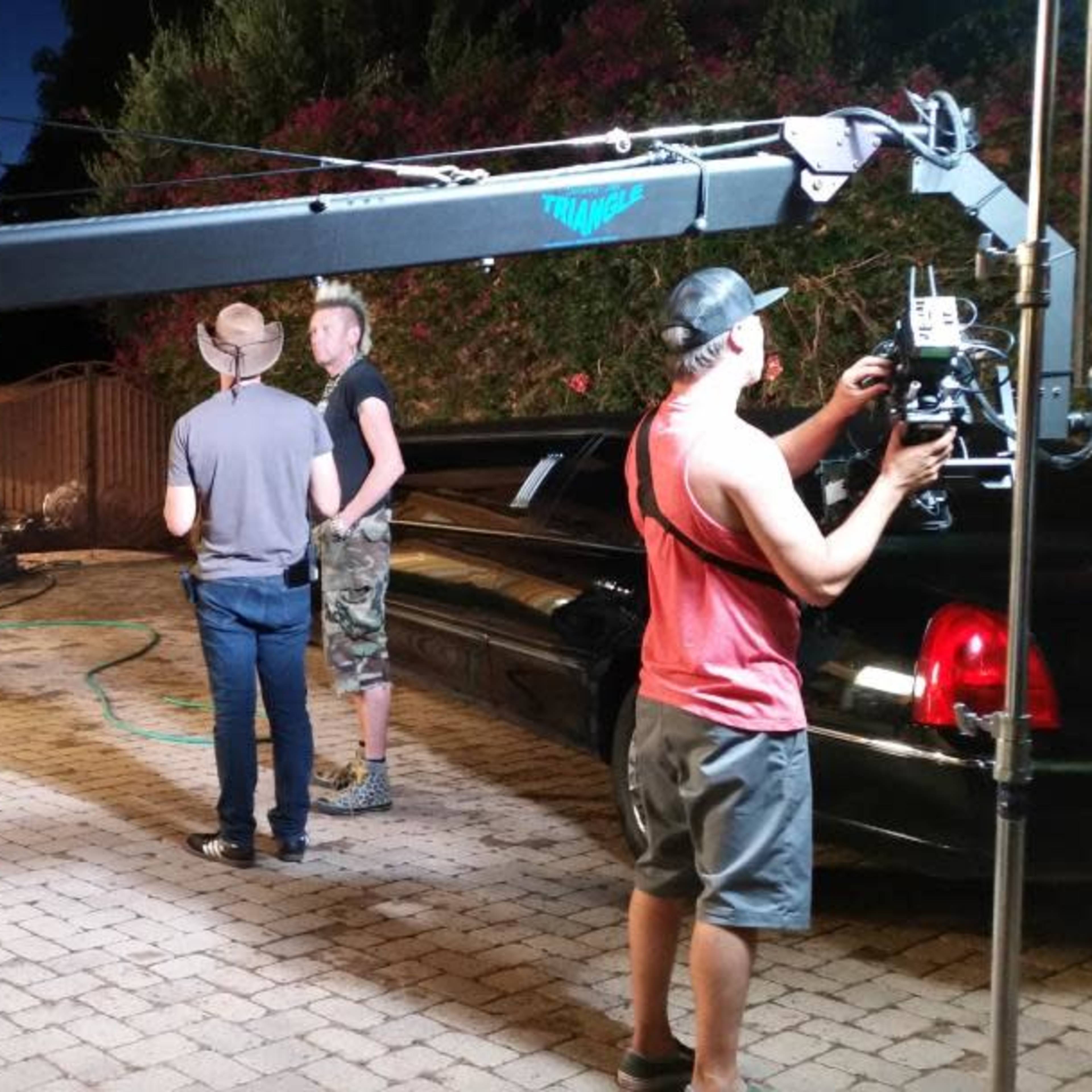 A film crew sets up equipment on a driveway outside a large house while performers interact nearby.