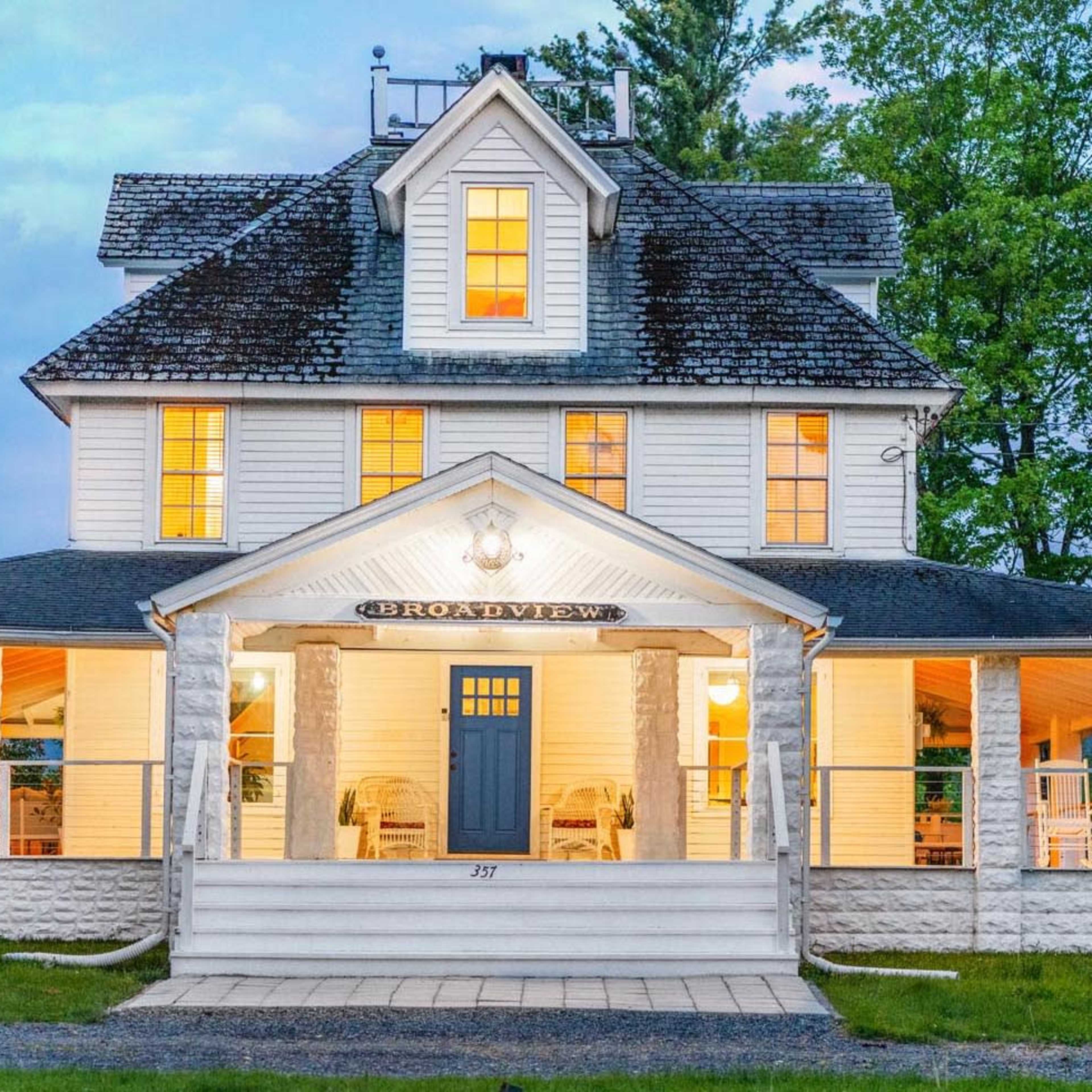 A large, white two-story house with a covered porch, surrounded by trees and illuminated windows.