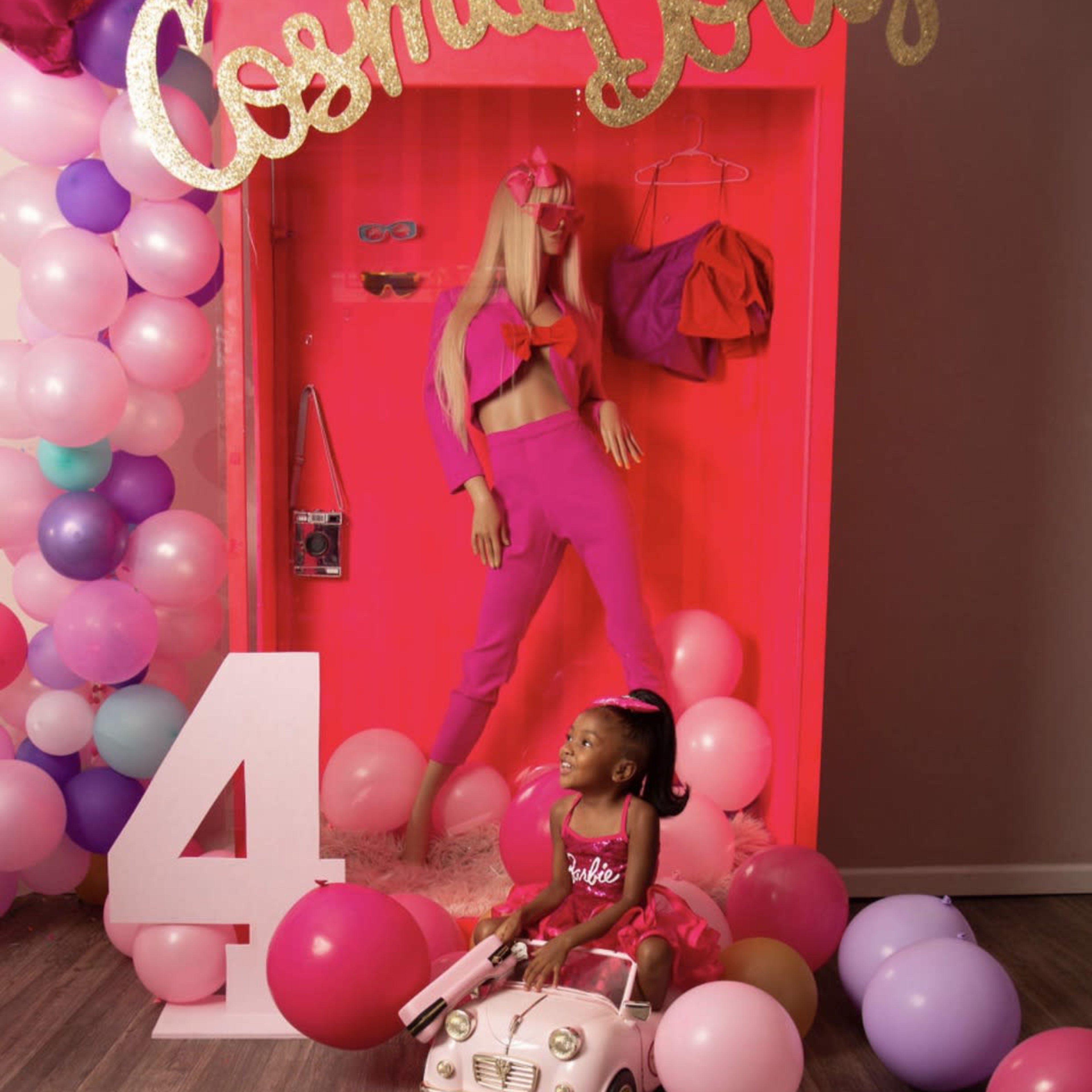 A young girl in a pink dress sits in a toy car surrounded by colorful balloons and a mannequin dressed in pink, with the words "Cosmo Dolly" displayed above.
