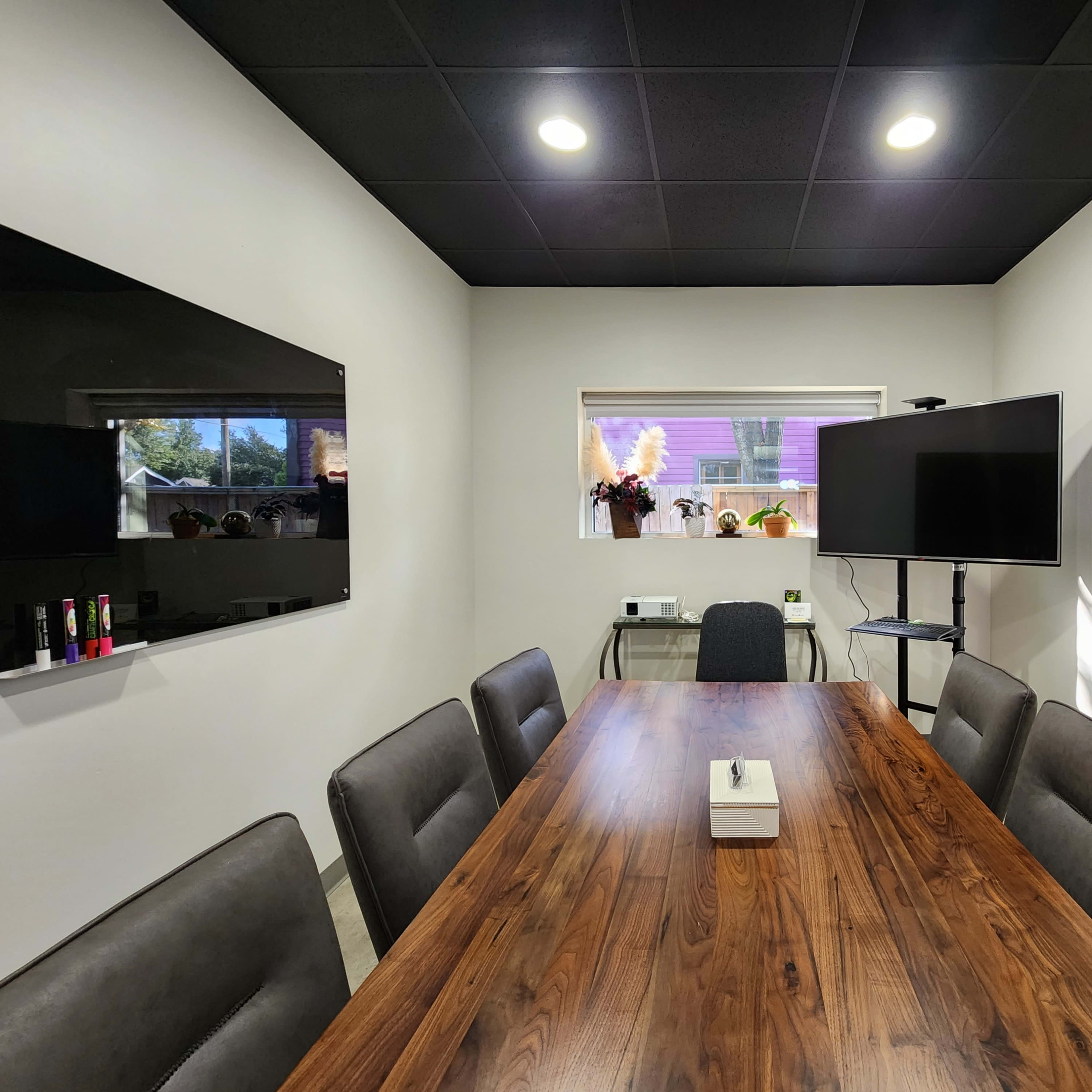 A conference room features a long wooden table surrounded by eight chairs, with two televisions mounted on the walls and a window offering natural light.