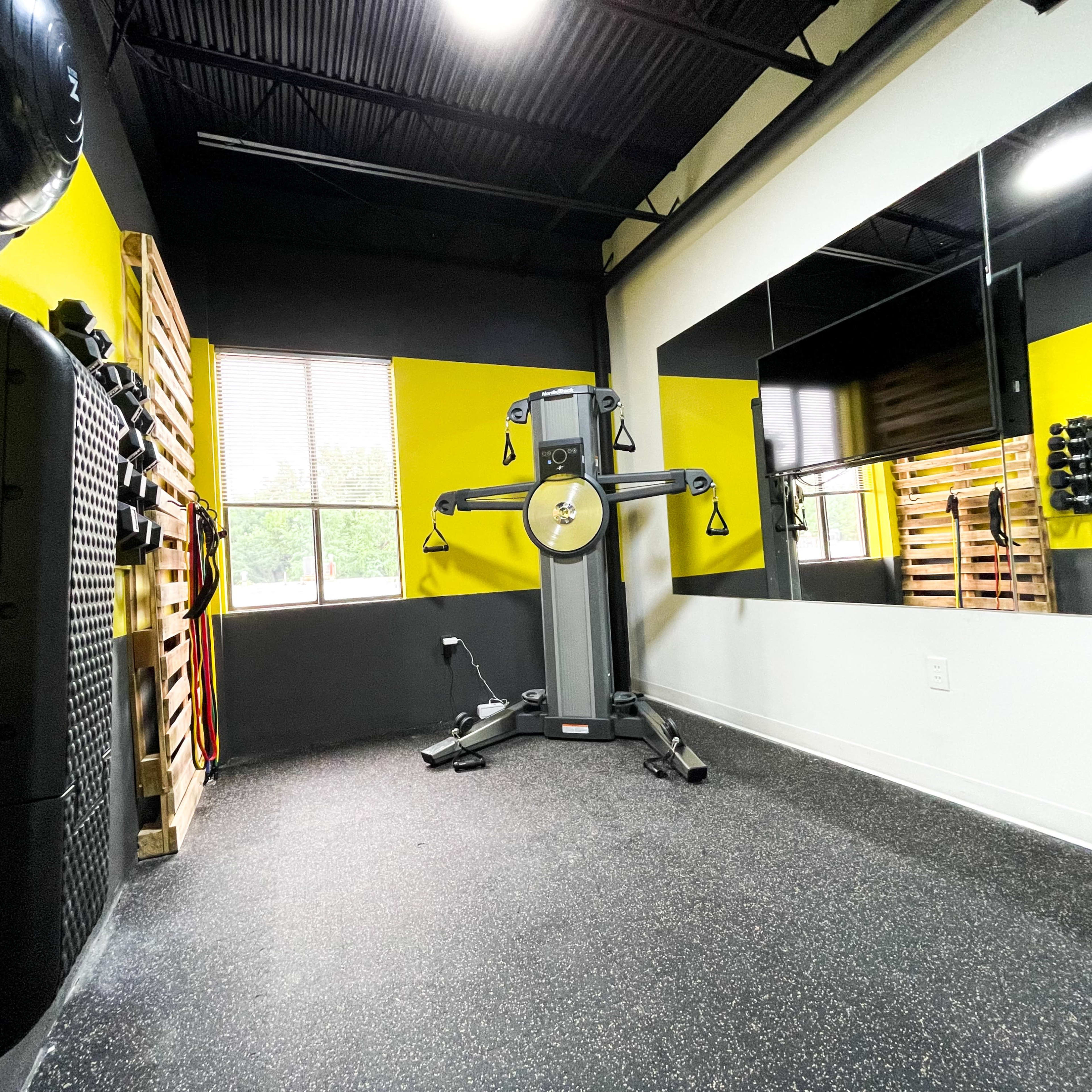 A fitness room equipped with exercise machines, free weights, and a large mirror, featuring black and yellow walls and a rubber floor.