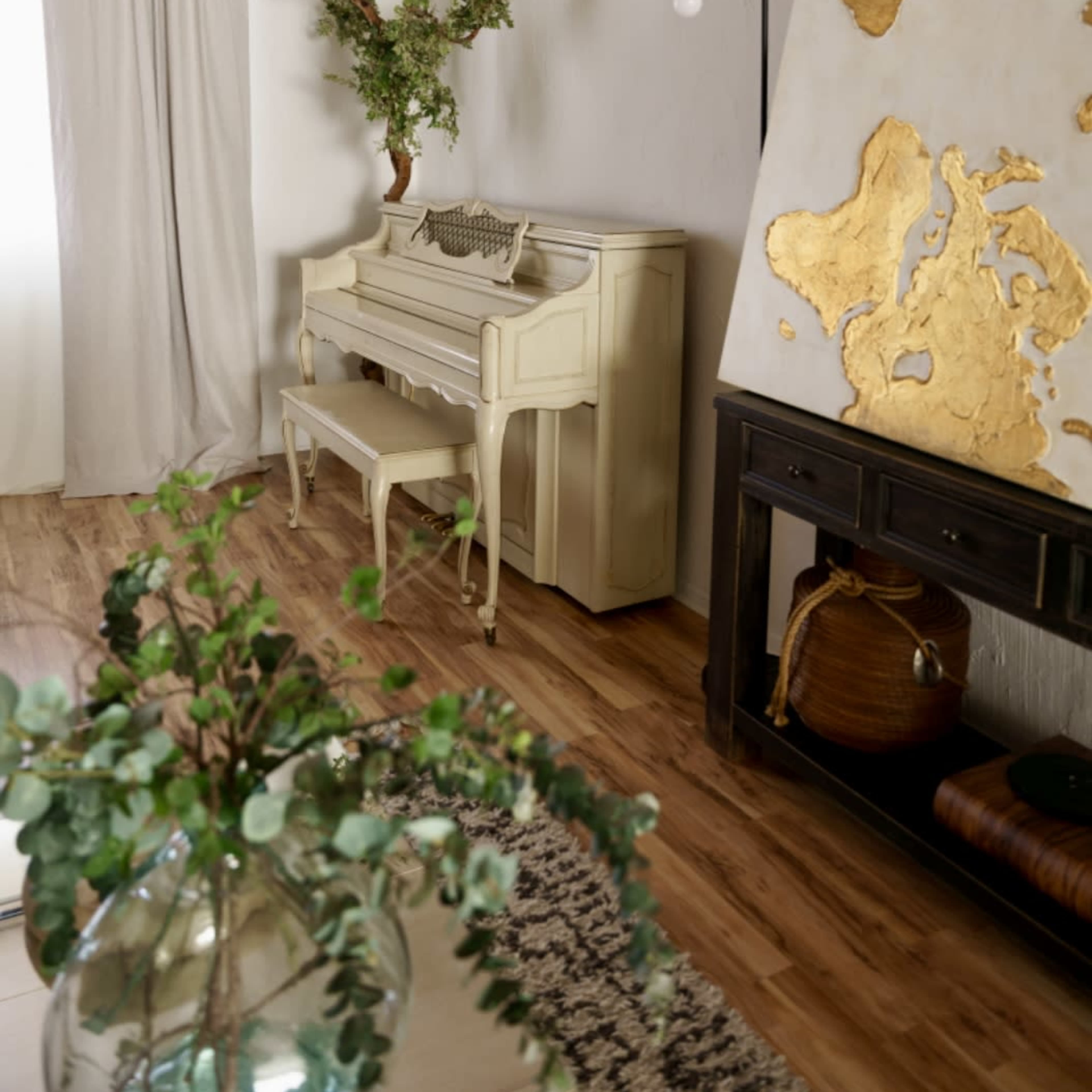 A vintage piano is situated beside a wall featuring a large, gold-leaf world map, with a decorative plant placed above it in a neutral-colored room.