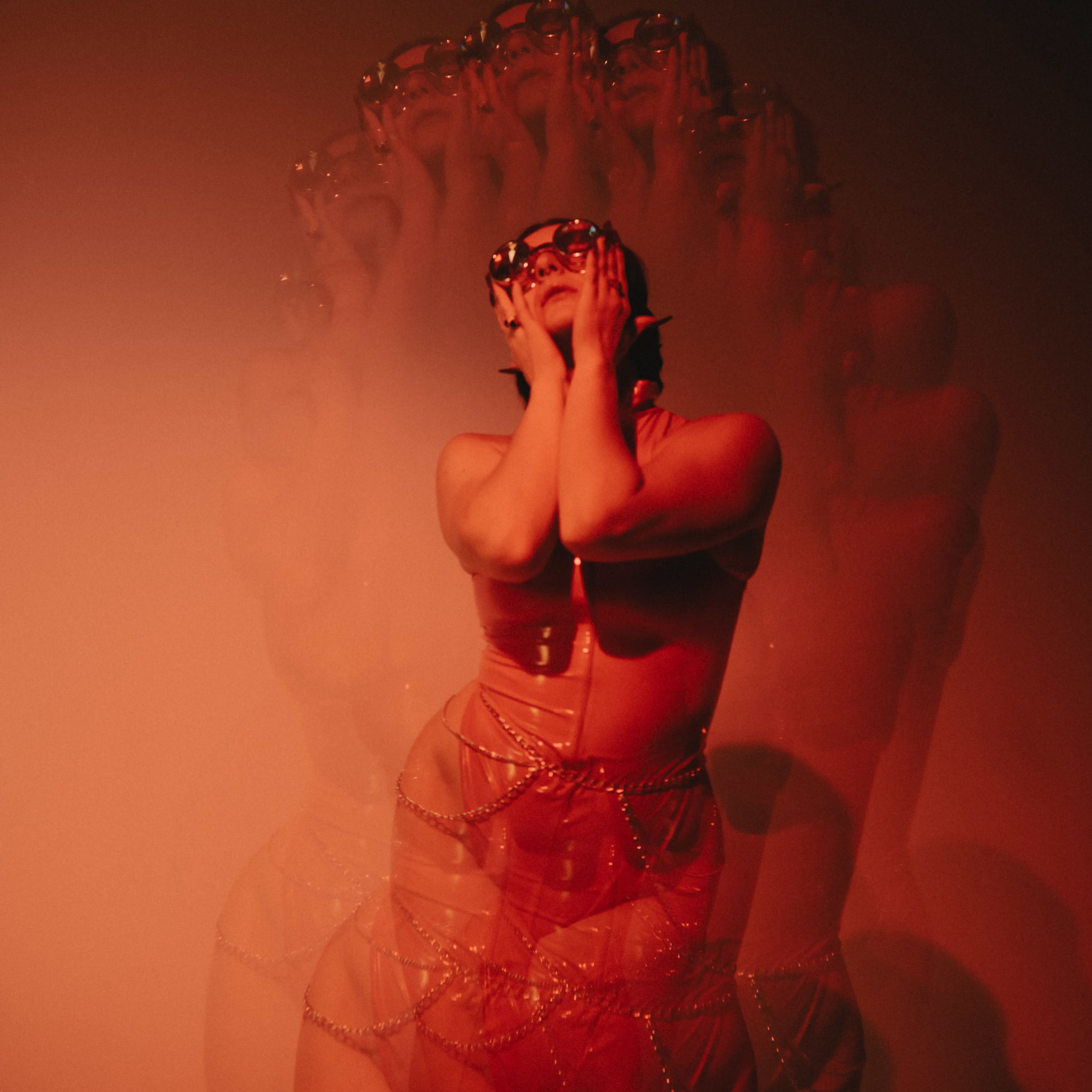 A figure wearing sunglasses poses against a softly lit background, creating a blurred effect that suggests movement.