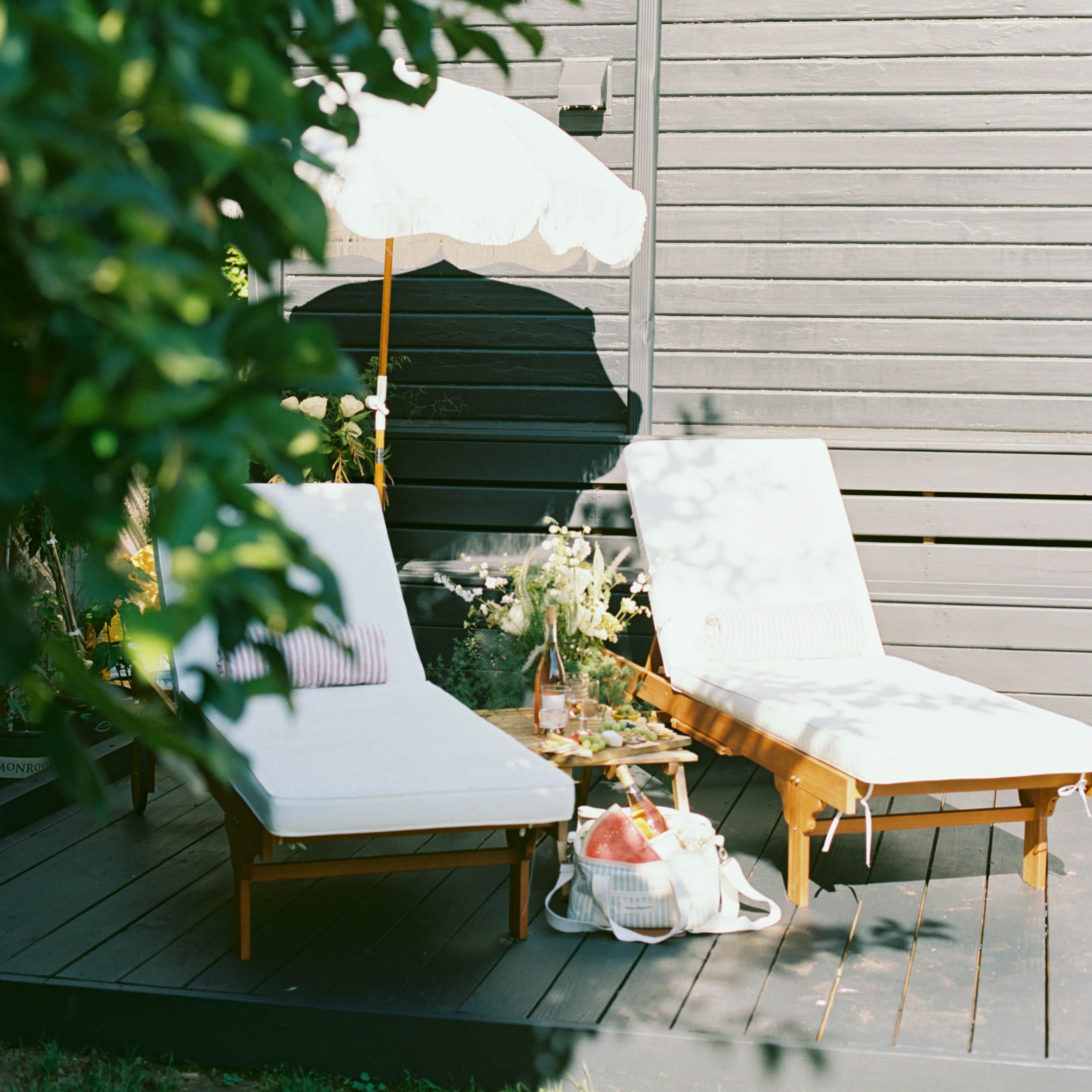 Two lounge chairs with a small umbrella are positioned on a deck next to a potted plant and a basket of apples.