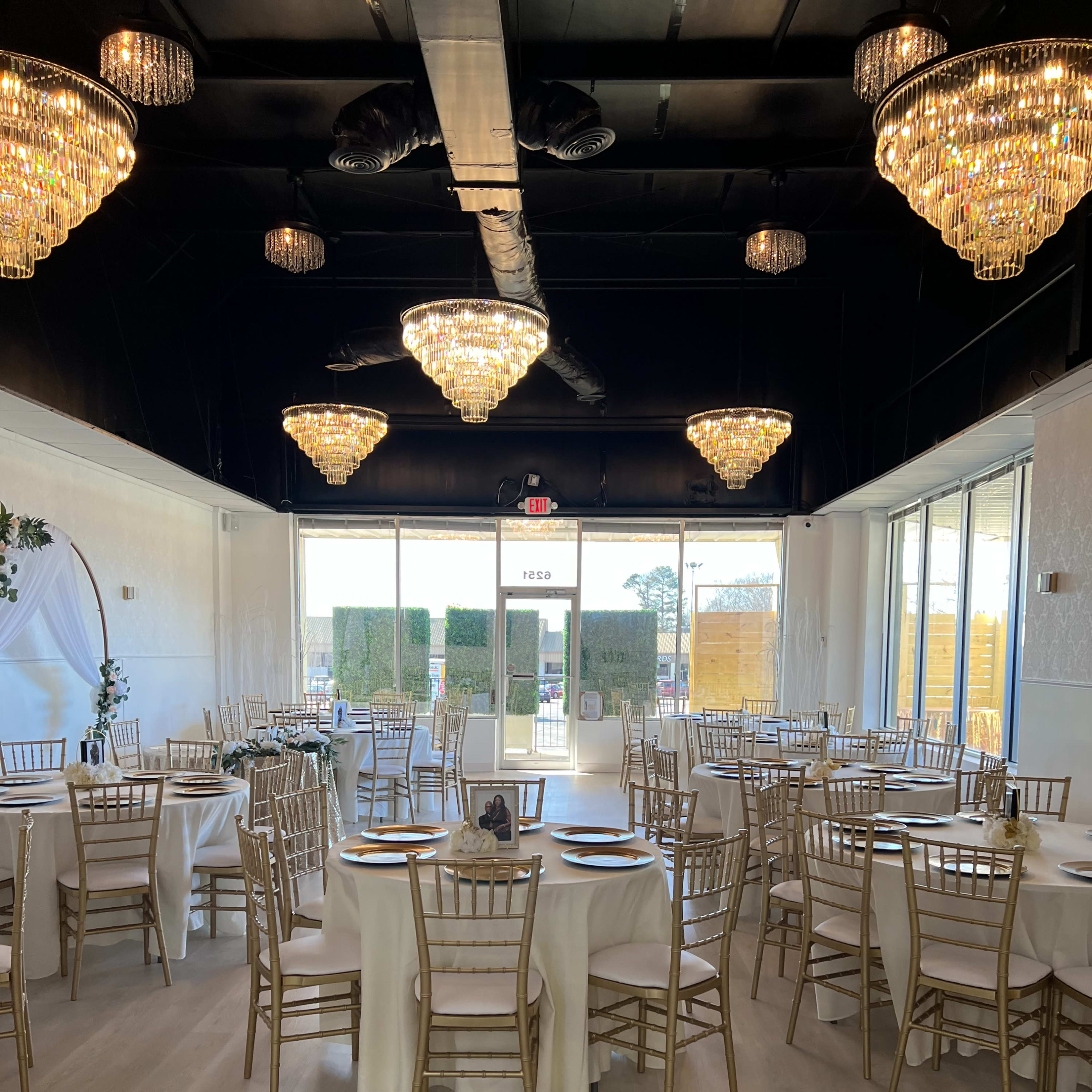 A well-lit banquet hall with gold-accented tables set for a formal event, featuring chandeliers and large windows inviting natural light.