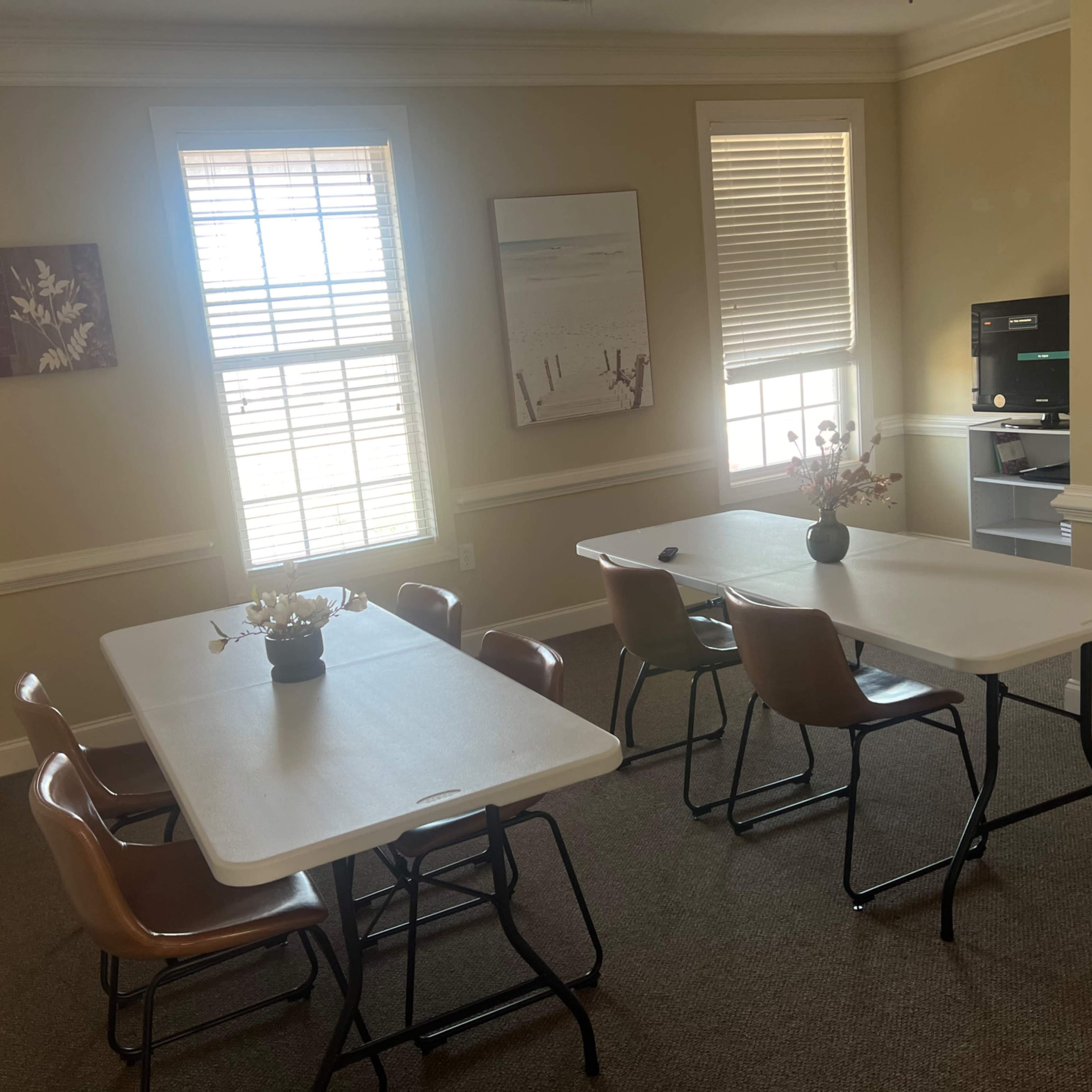 A small meeting room with two foldable tables and several chairs, featuring a flower centerpiece and a large window with blinds.
