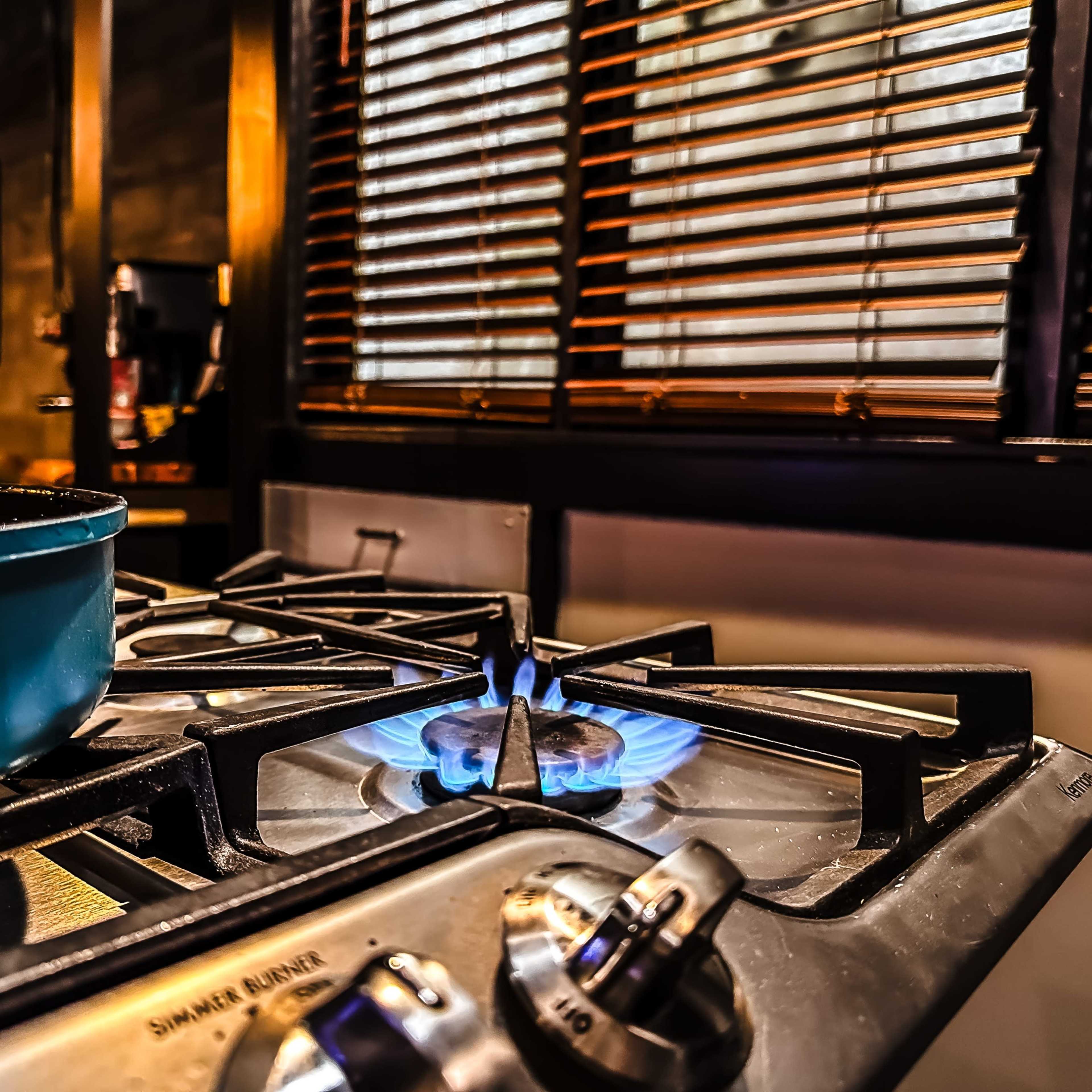 A blue flame burns on a gas stove with a cooking pan nearby, set against a backdrop of wooden blinds.