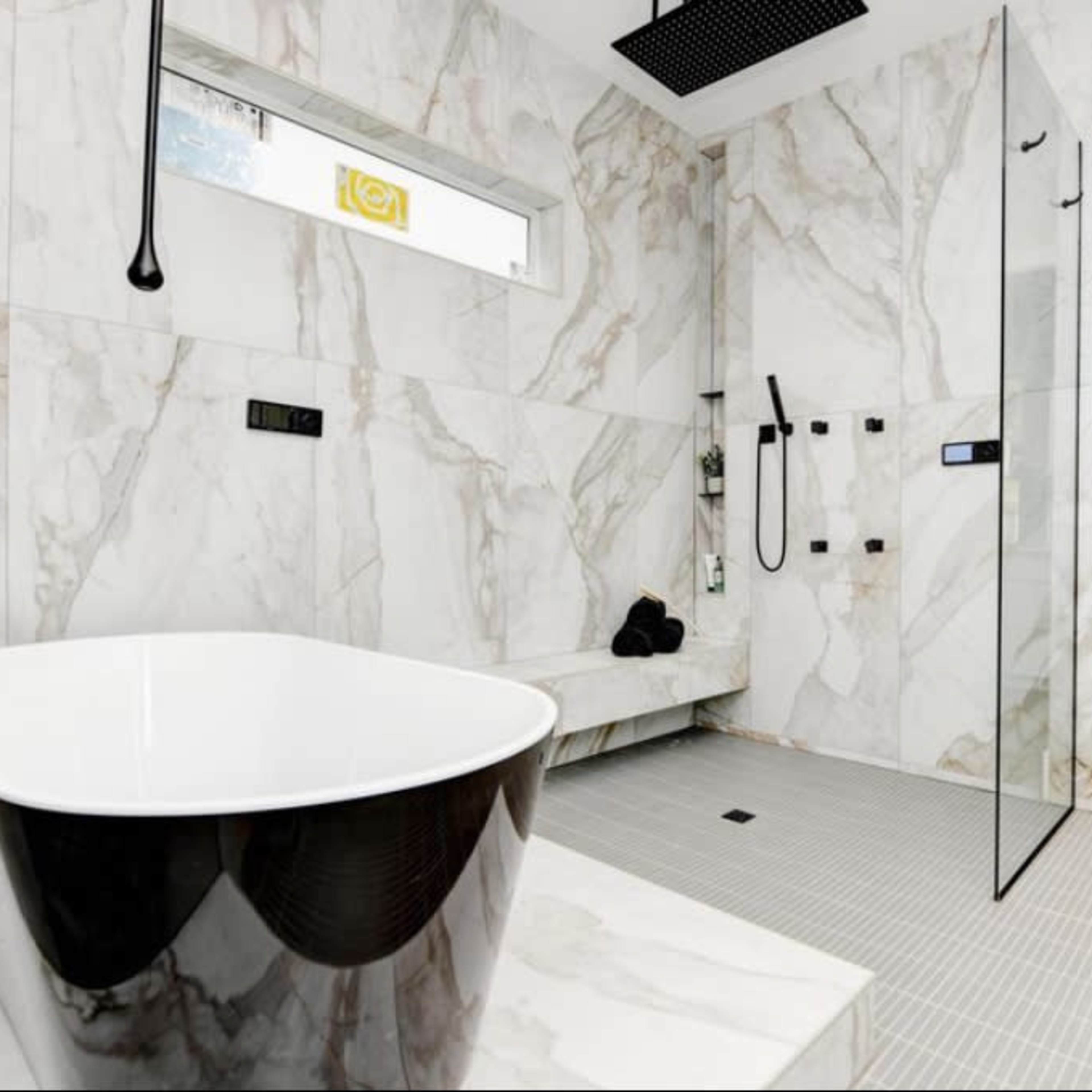 A modern bathroom featuring a freestanding black and white bathtub, a glass shower enclosure, and marble walls with a gray tiled floor.