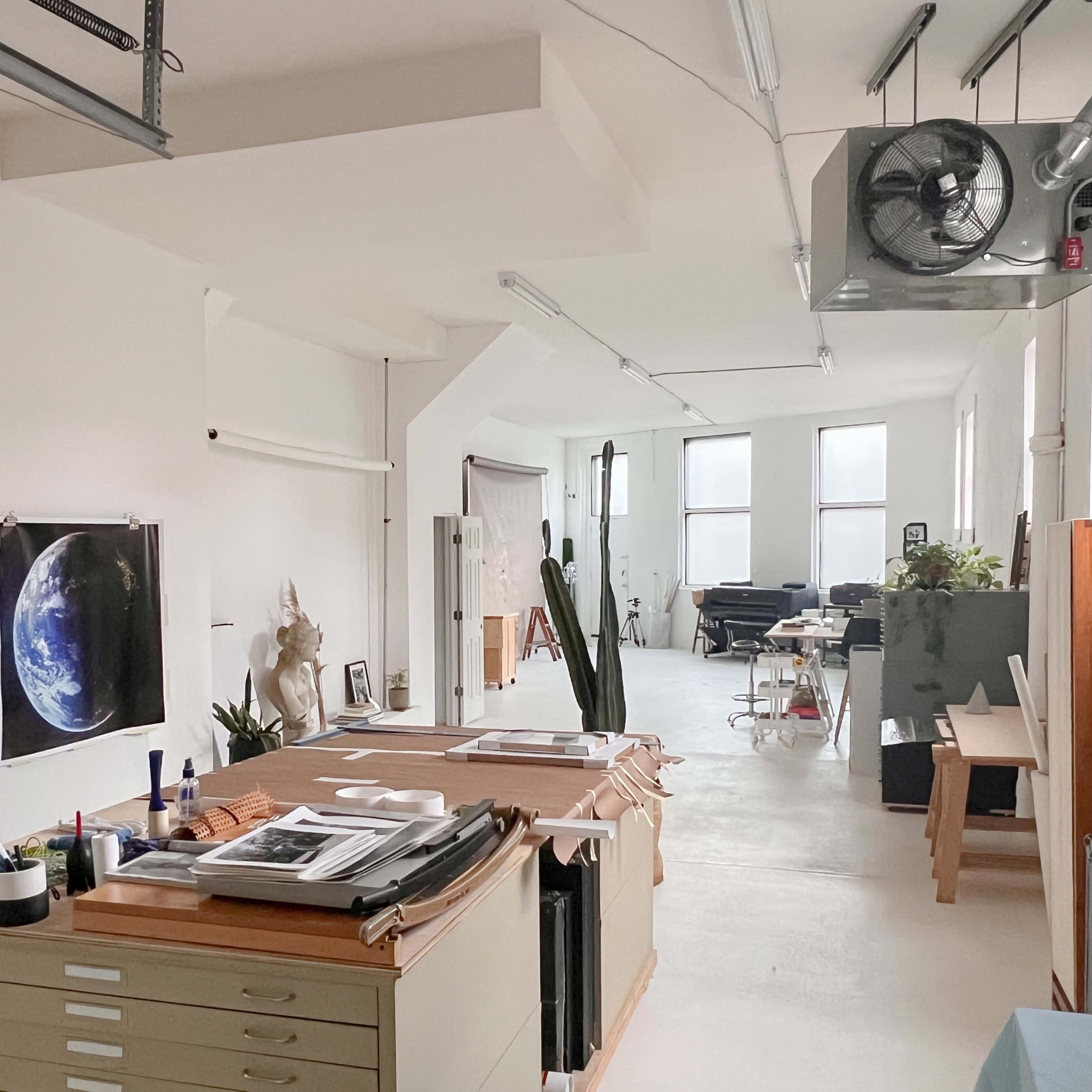 A bright art studio with several worktables, an assortment of artwork on the walls, and a view of additional creative spaces in the background.