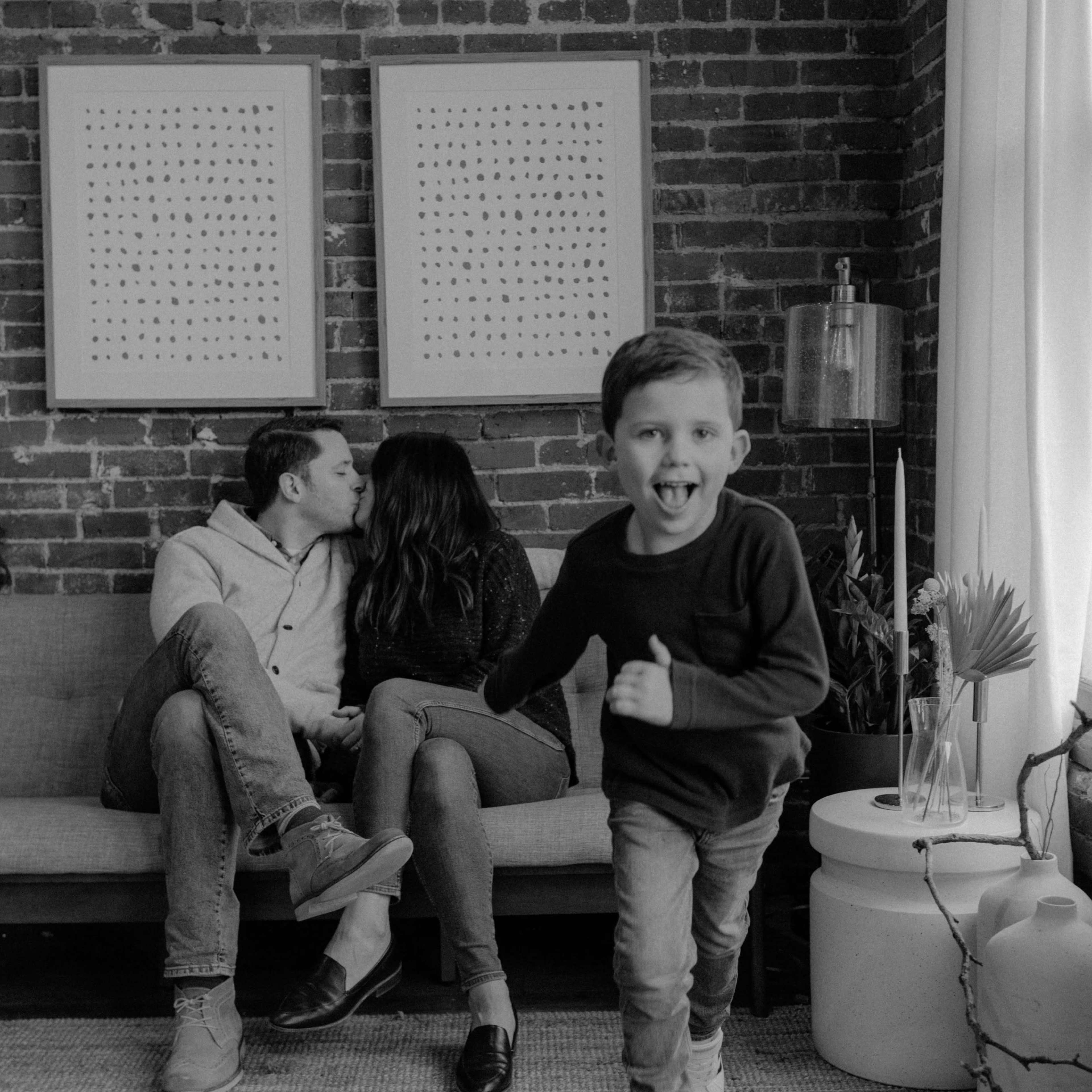 A child runs playfully in the foreground while a couple shares a kiss on a sofa against a brick wall adorned with framed art.