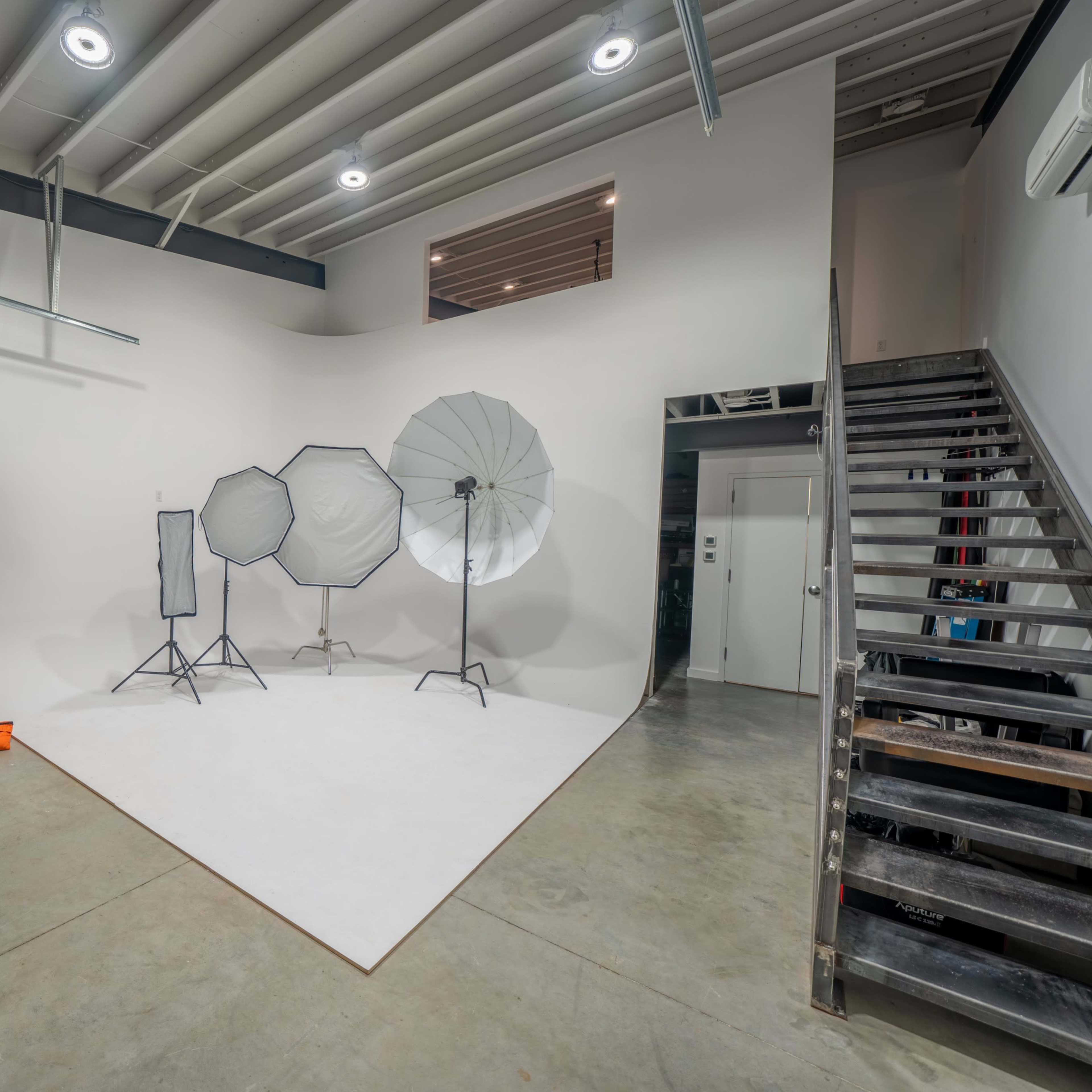 A photography studio features a backdrop, multiple light modifiers, a ladder, a TV screen, and a staircase.
