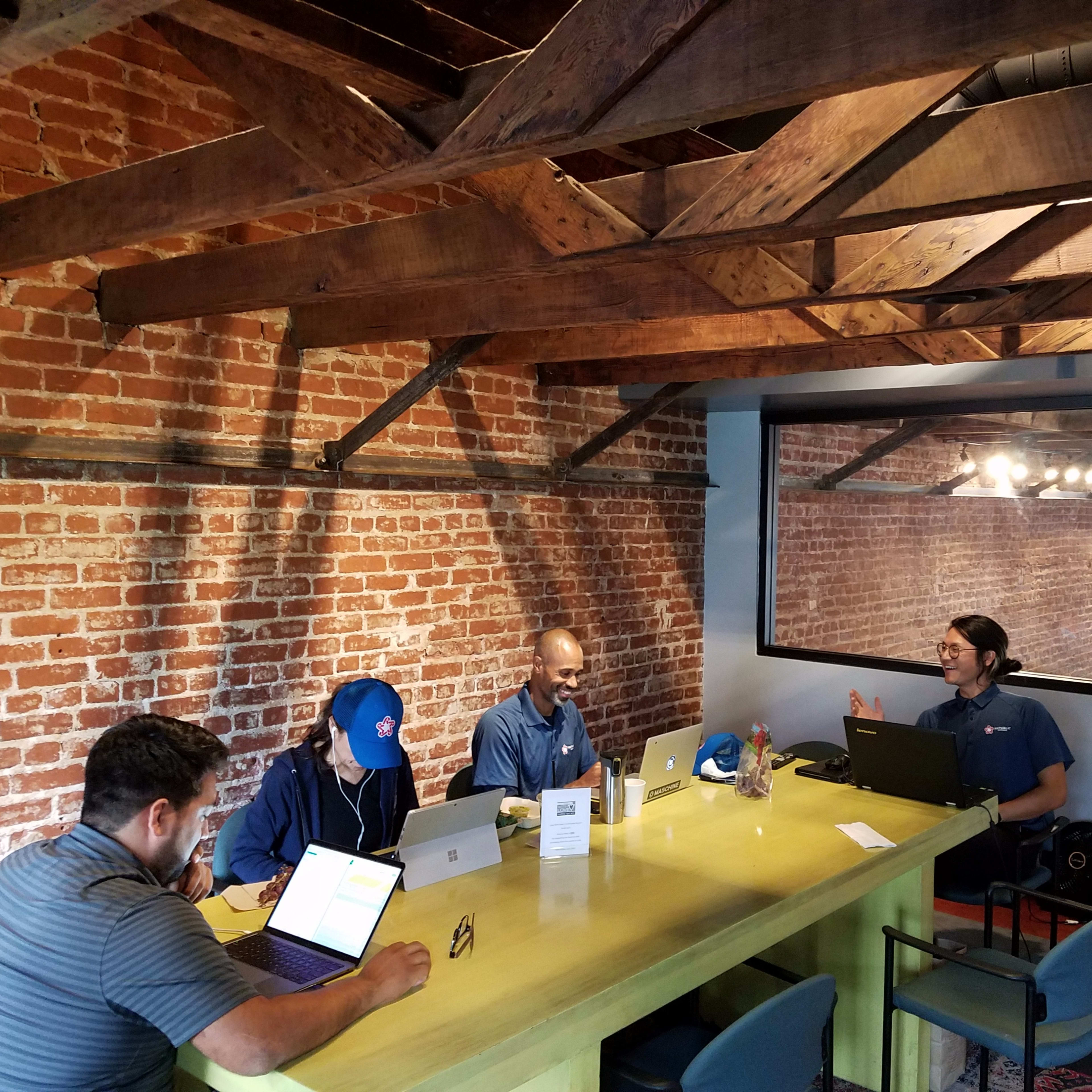 A group of four people work on laptops around a green table in a room with exposed brick walls and wooden beams.