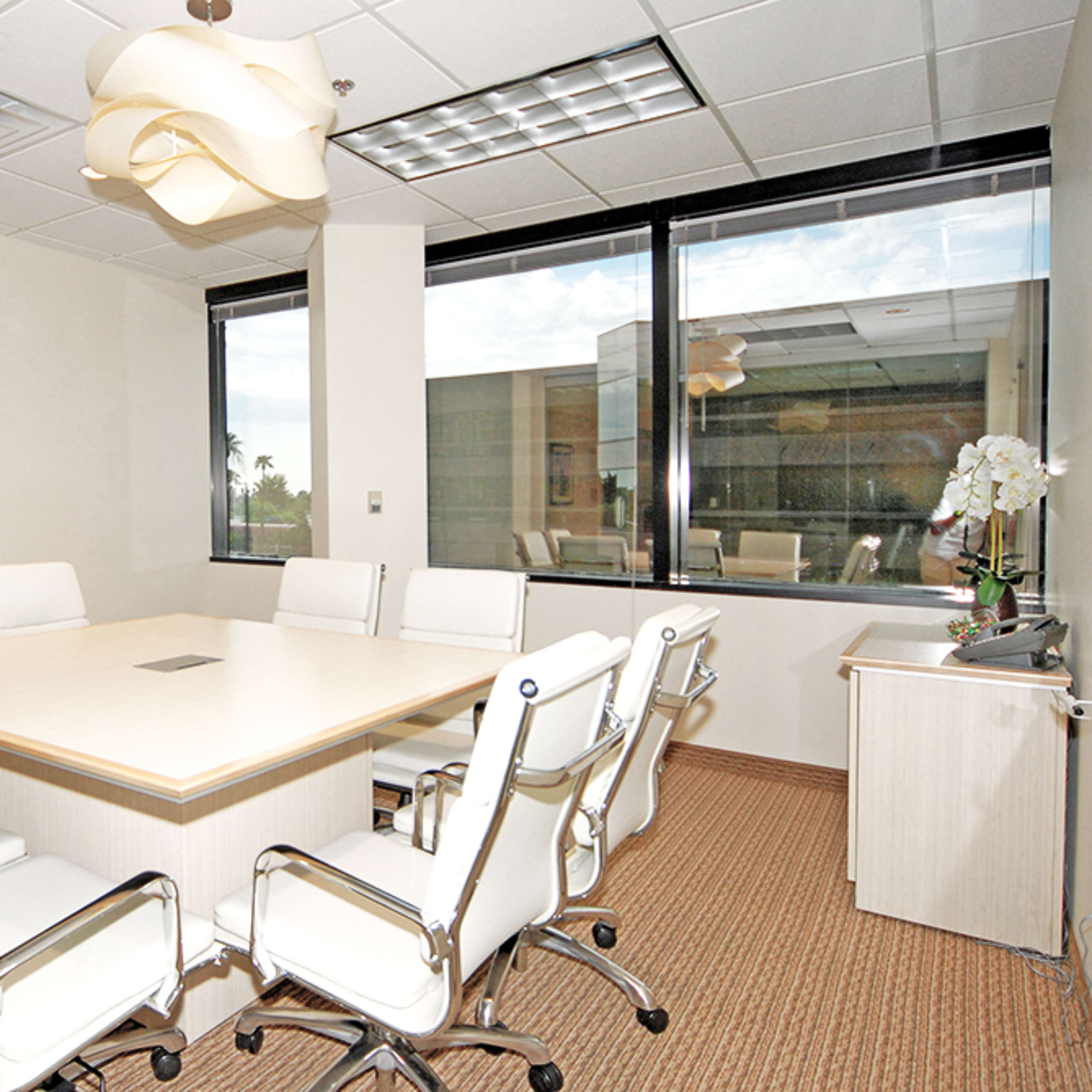 A modern conference room features a large wooden table surrounded by white chairs, with a television mounted on one wall and large windows letting in natural light.