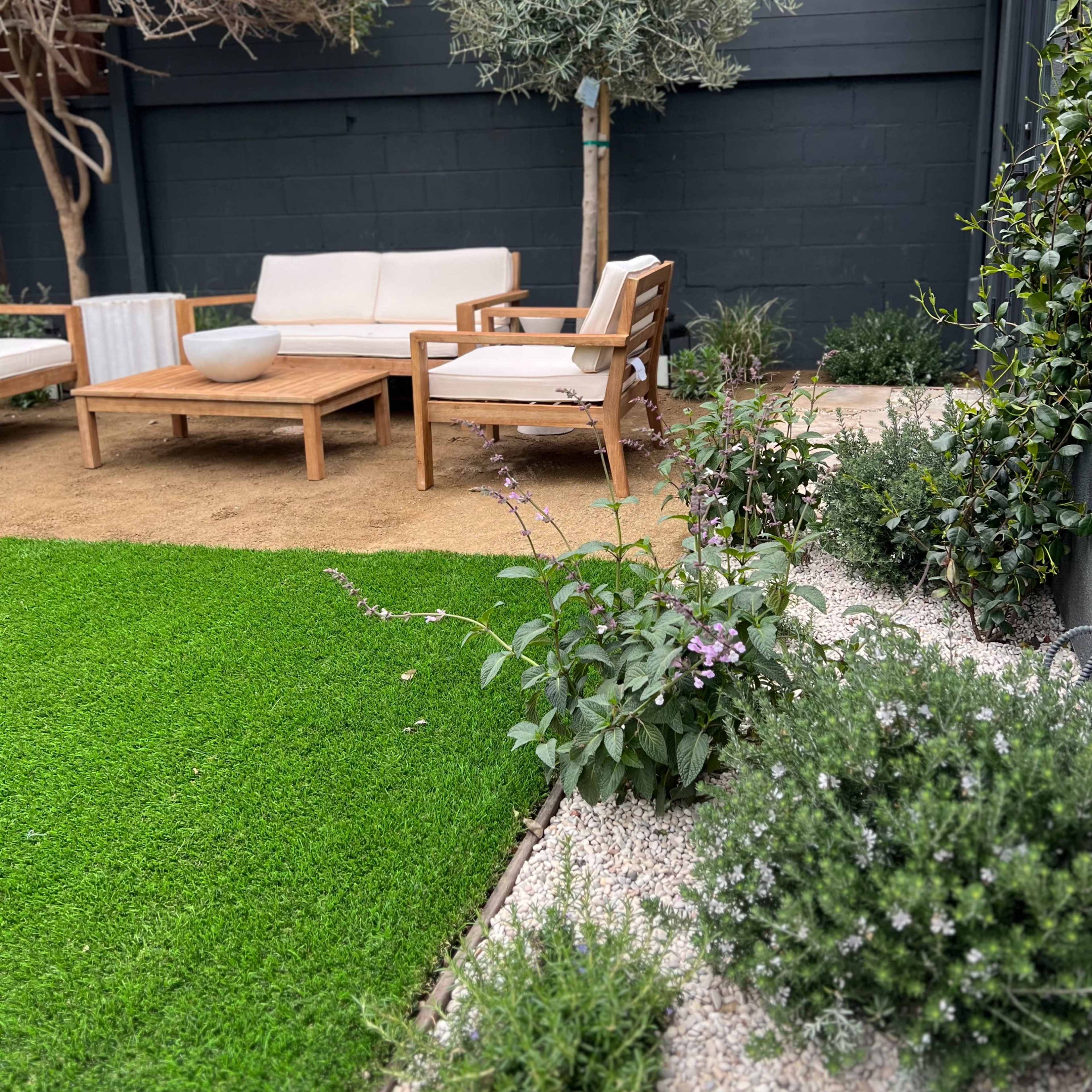 A landscaped garden area with a sitting arrangement featuring wooden chairs and a table, surrounded by green grass and gravel pathways, along with various plants and a small tree.