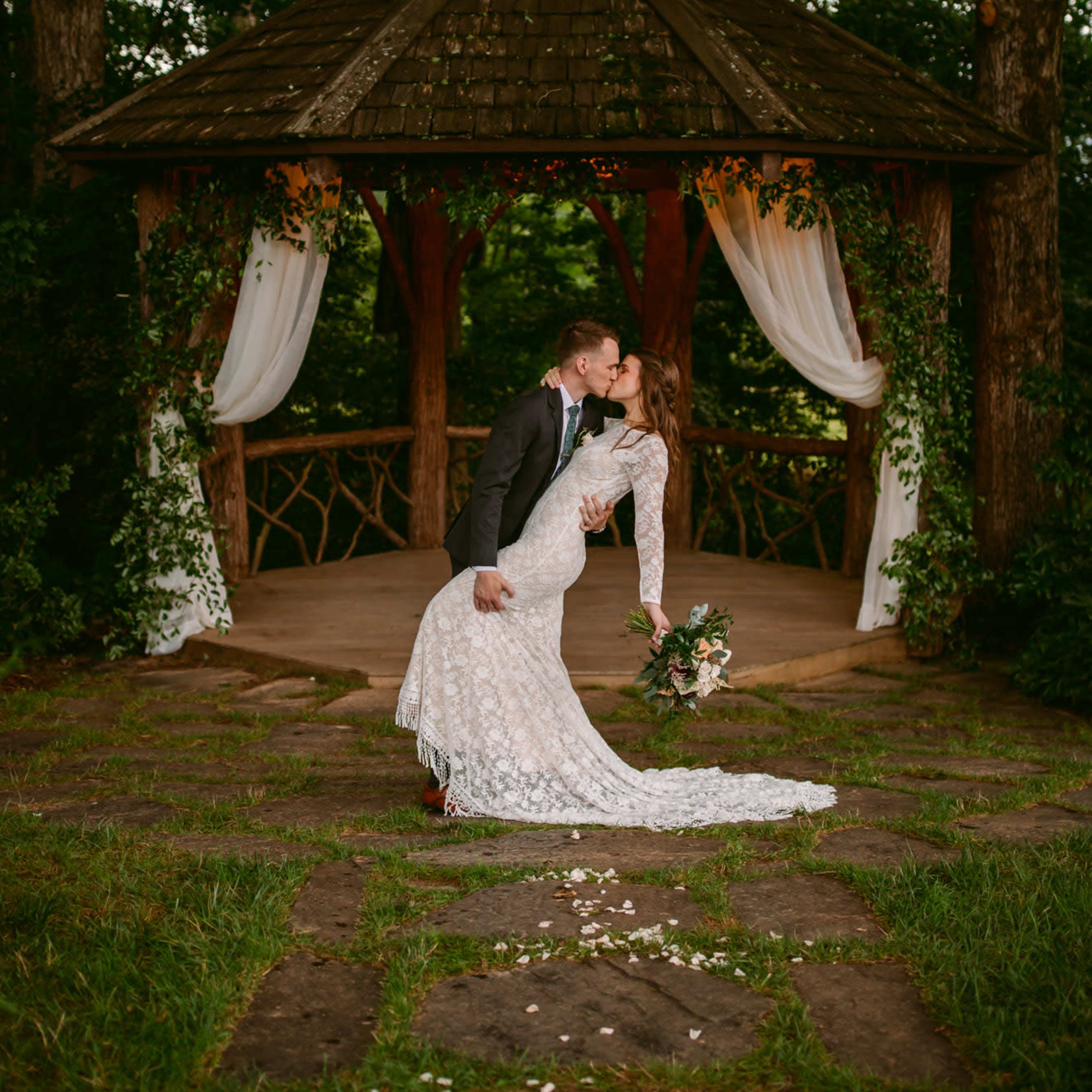 A couple in wedding attire shares a kiss under a gazebo surrounded by trees.