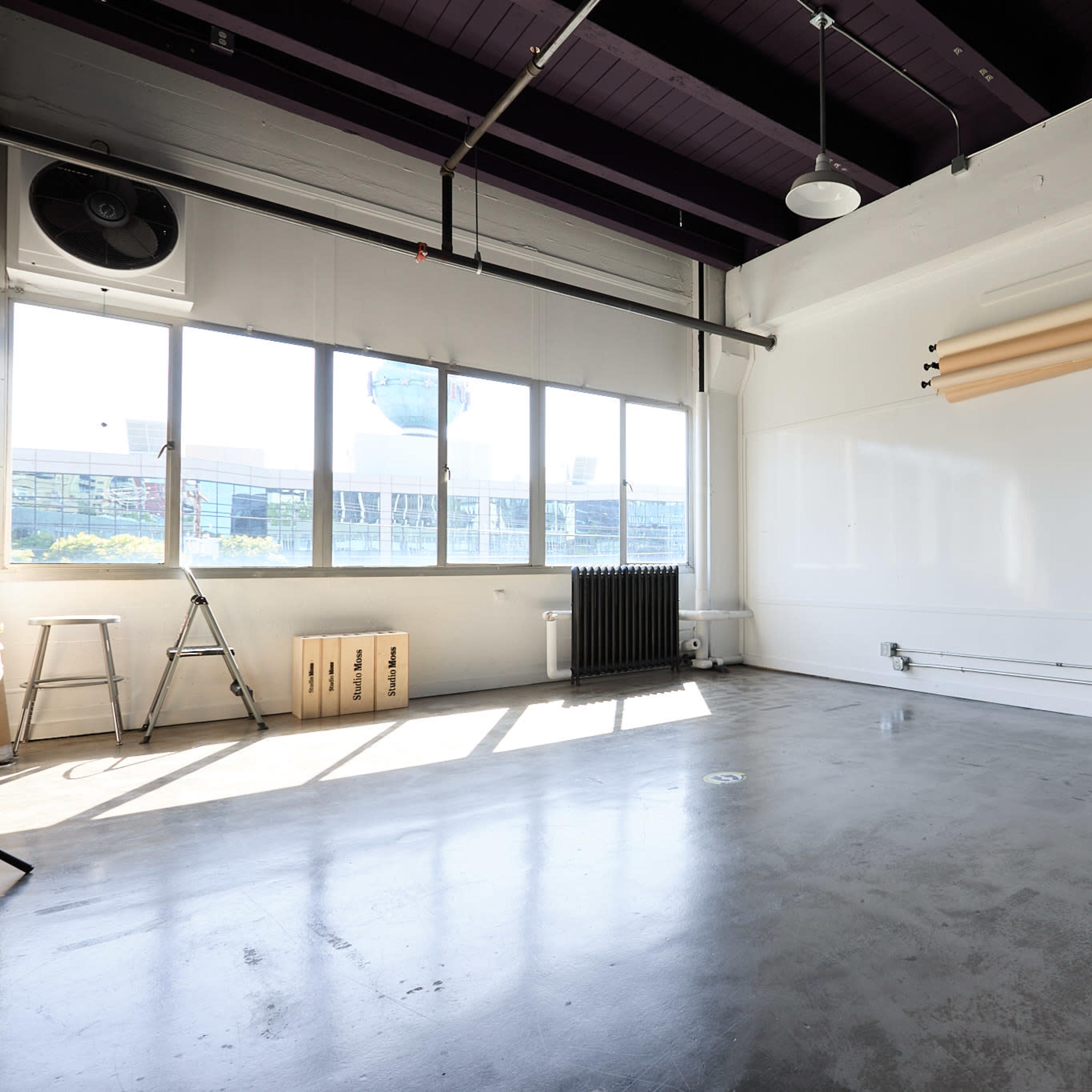 A spacious, minimalistic studio with large windows, concrete flooring, and a few pieces of furniture, including a cart and metal stools.